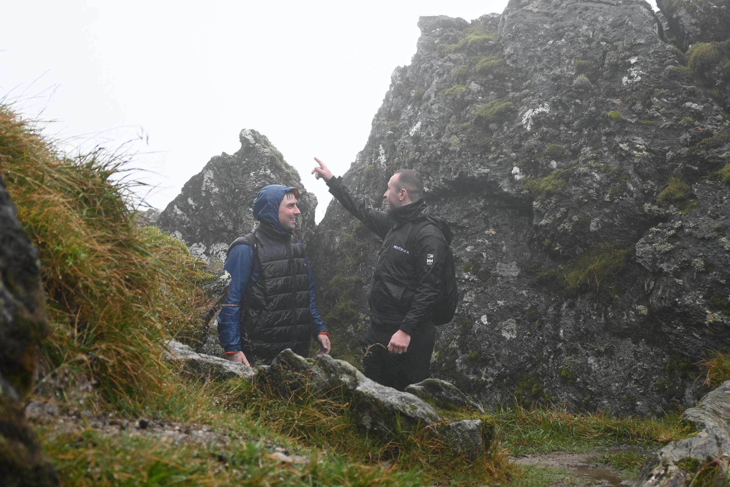 Two men hiking and talking among rocky terrain with moss and grass, surrounded by foggy weather.