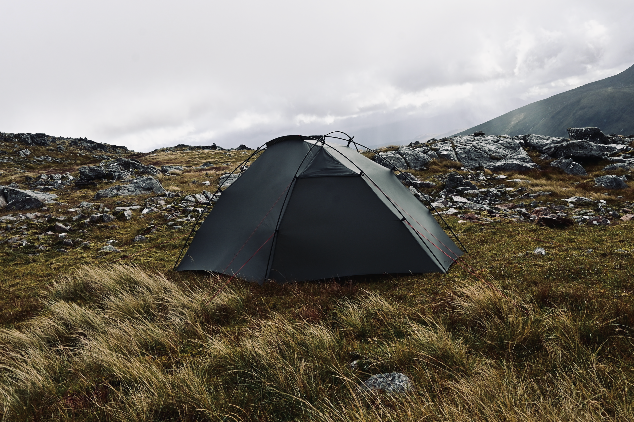 A tent set up on a rocky hillside with grass and large rocks, mountainous terrain, and cloudy sky in the background.