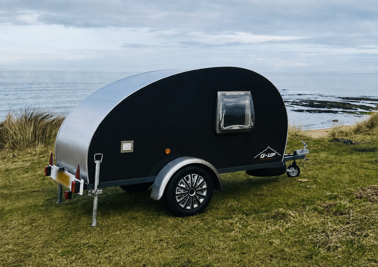 Black teardrop-shaped camper trailer with a silver front panel, positioned on grassy coast with ocean in the background. The camper has a small window, a wheel, and a hitch for towing.