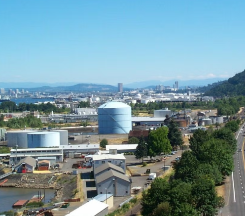 Industrial area with storage tanks, warehouses, trees, and a highway with cars in the foreground, and a city skyline with high-rise buildings in the background under a blue sky.