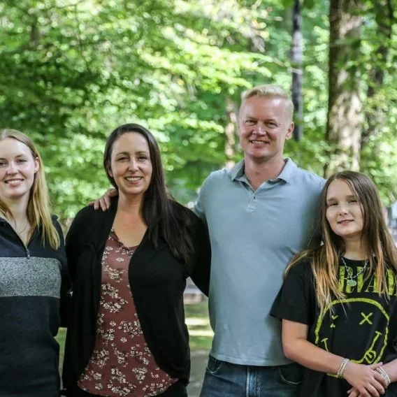 Four people, two women and two teenagers, standing outdoors in front of a green, leafy background, smiling for the camera.