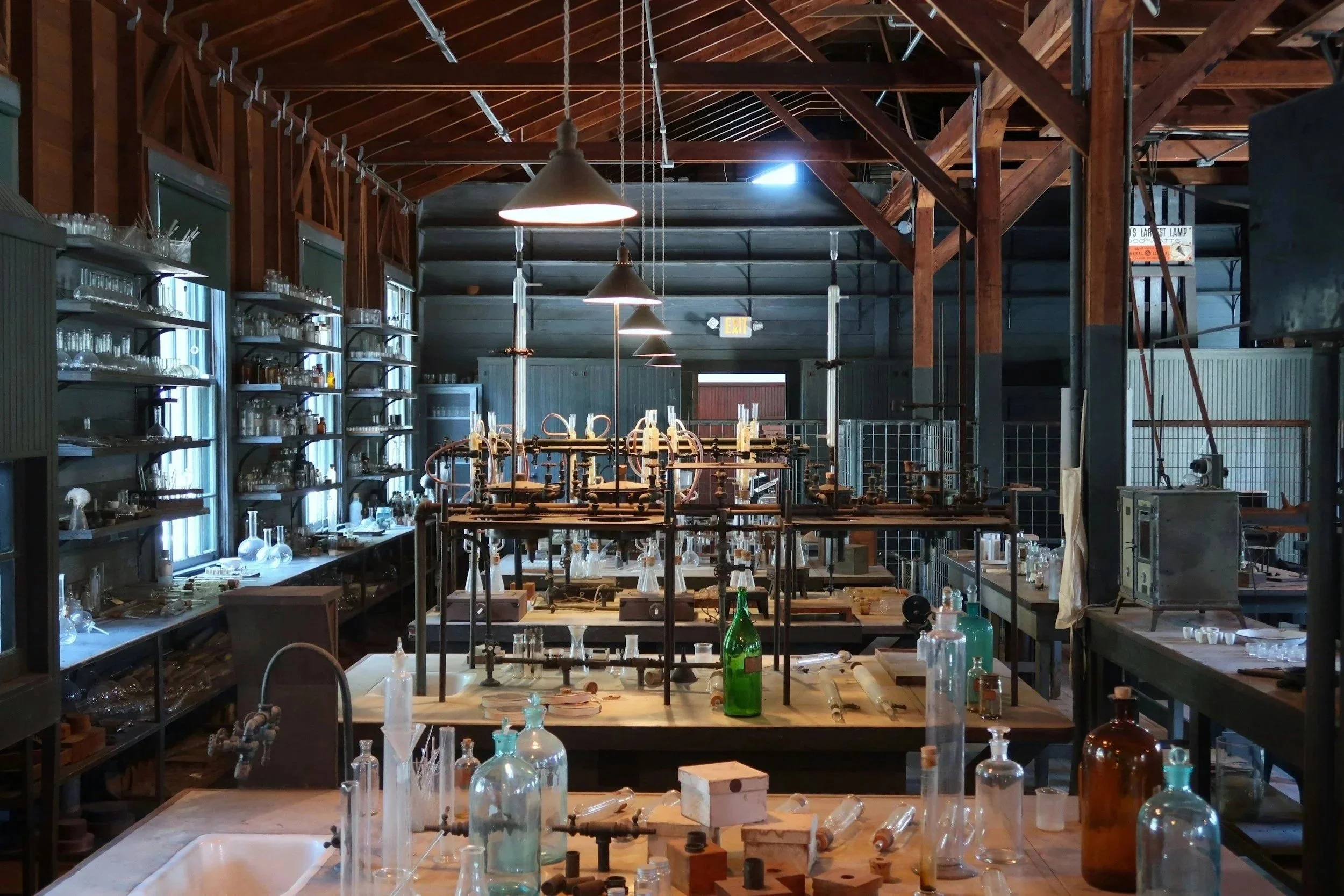 Intérieur d'un laboratoire ou d'une salle de recherche avec des étagères remplies de bocaux en verre et de matériel scientifique, plusieurs lampes suspendues au plafond en bois, un grand plan de travail avec divers objets de laboratoire, et des murs en bois.