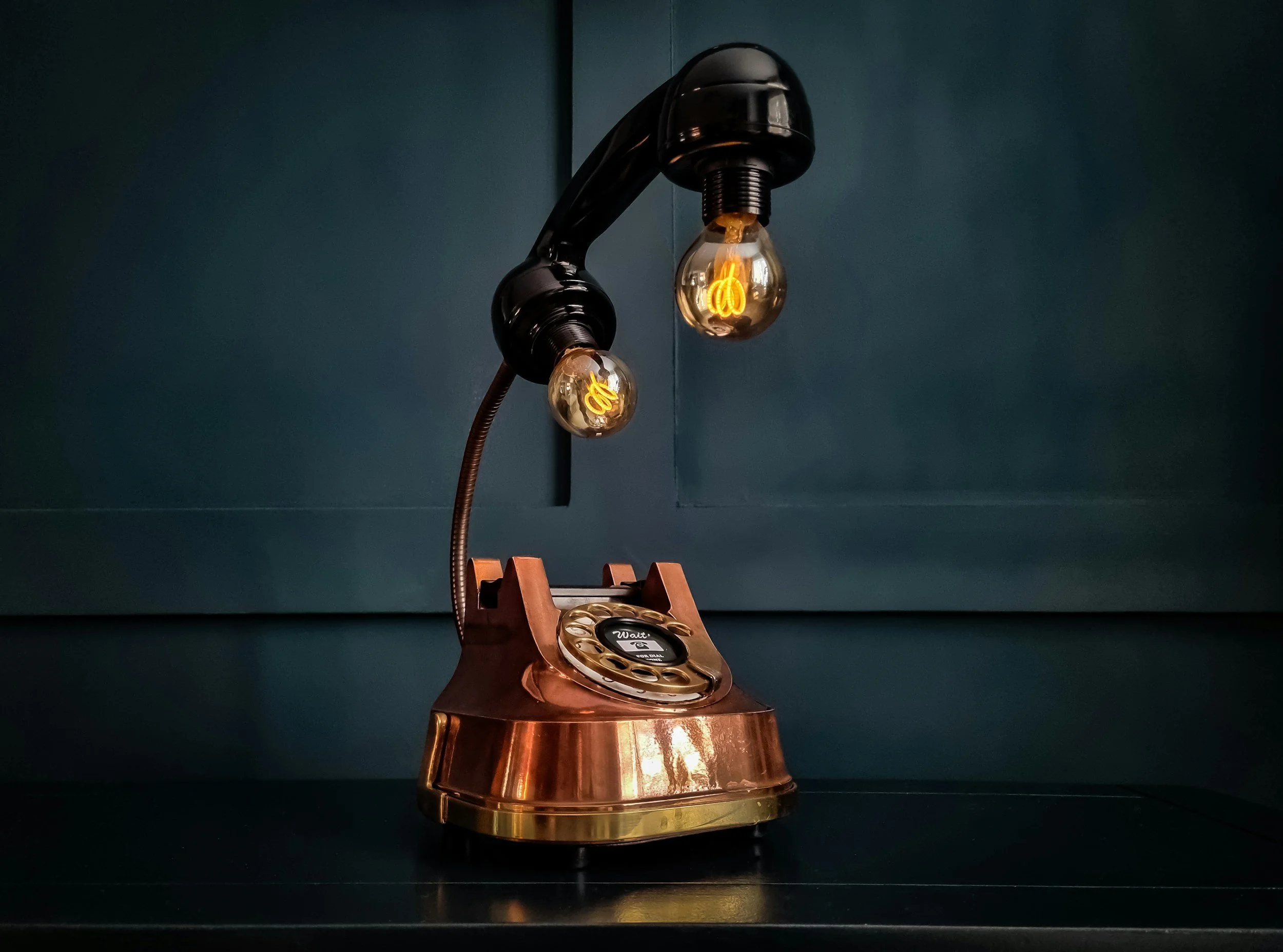 Lampe murale noire avec deux ampoules encastrées, une des ampoules allumée, en forme de filament décoratif, fixé à un mur foncé, avec un socle en cuivre.