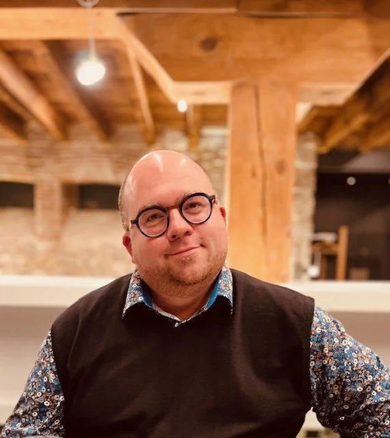 Un homme portant des lunettes, un pull noir et une chemise à motifs, dans une pièce avec une structure en bois visible au plafond.