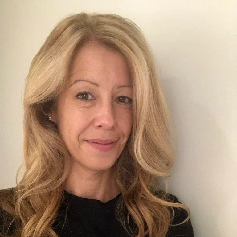 Femme avec longs cheveux blonds, souriant légèrement, portant une veste noire, debout devant un mur blanc.