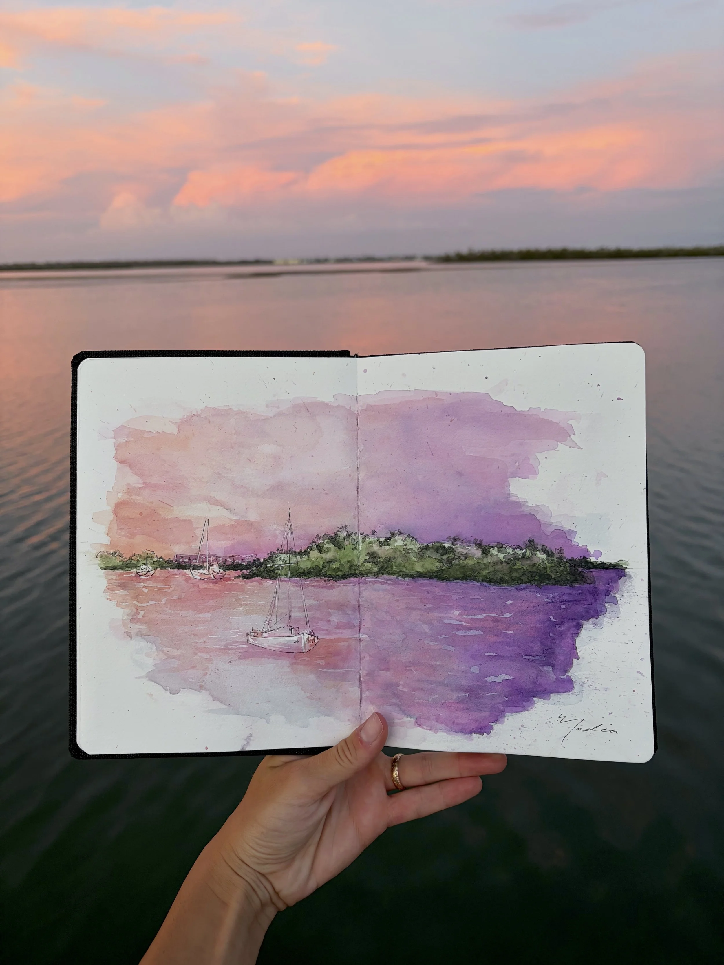 A hand holding a sketchbook with a watercolor painting of a boat and a landscape with water and trees, set against a sunset sky reflected on water.