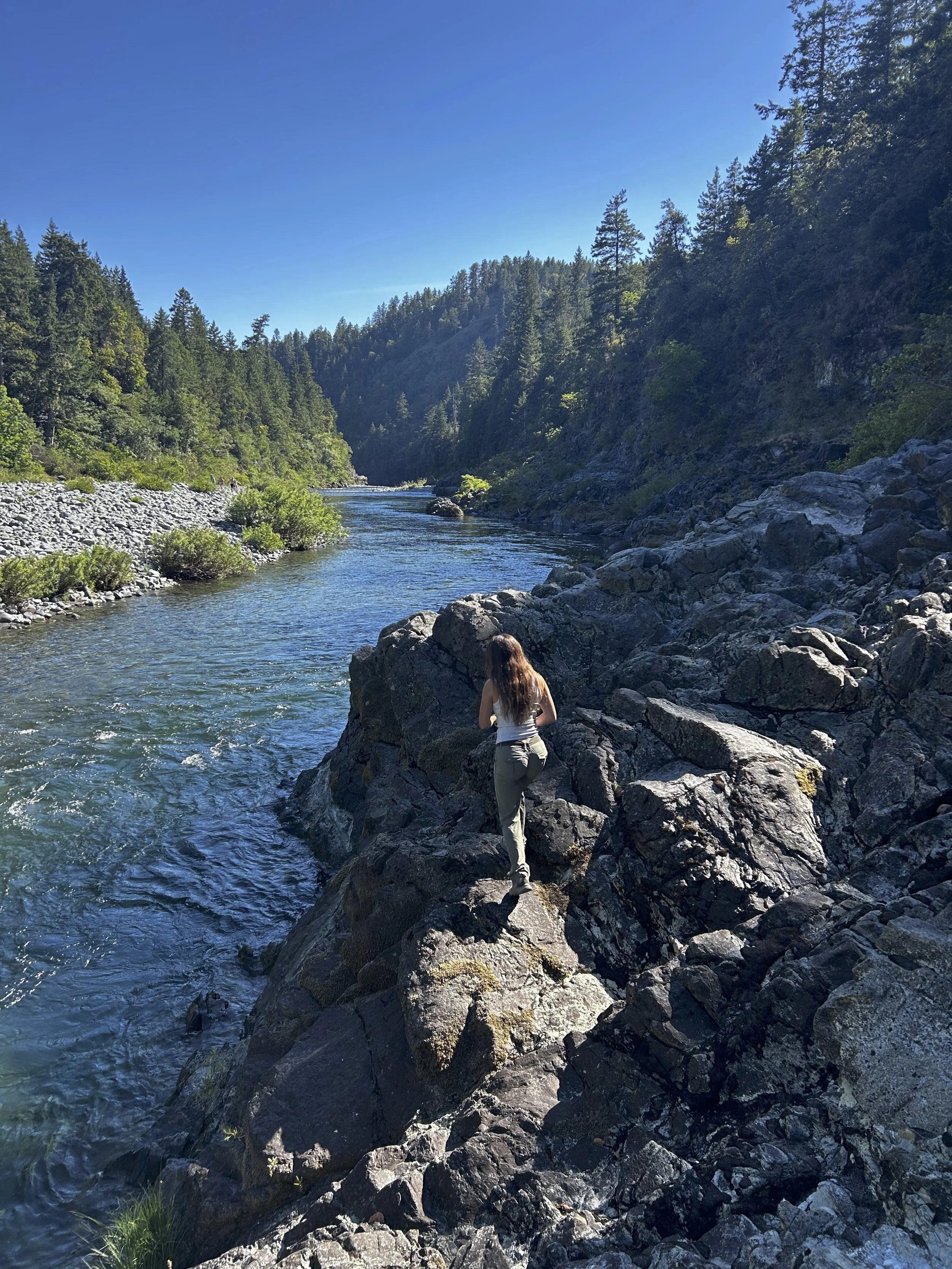 A woman standing on rocks beside a flowing river in a forested landscape under a clear blue sky.