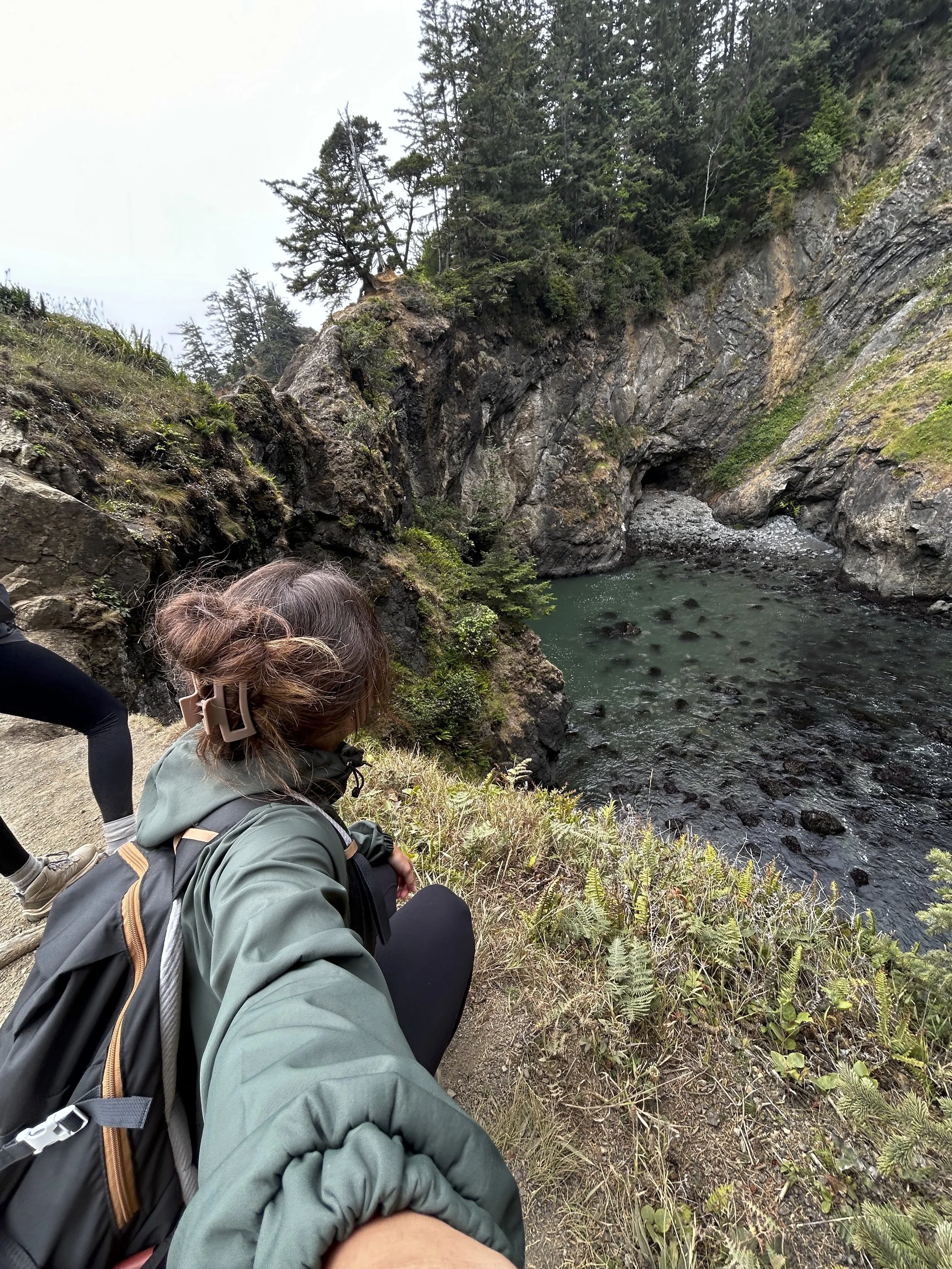 Hikers resting on a trail beside a river in a forested canyon with steep rocky walls.