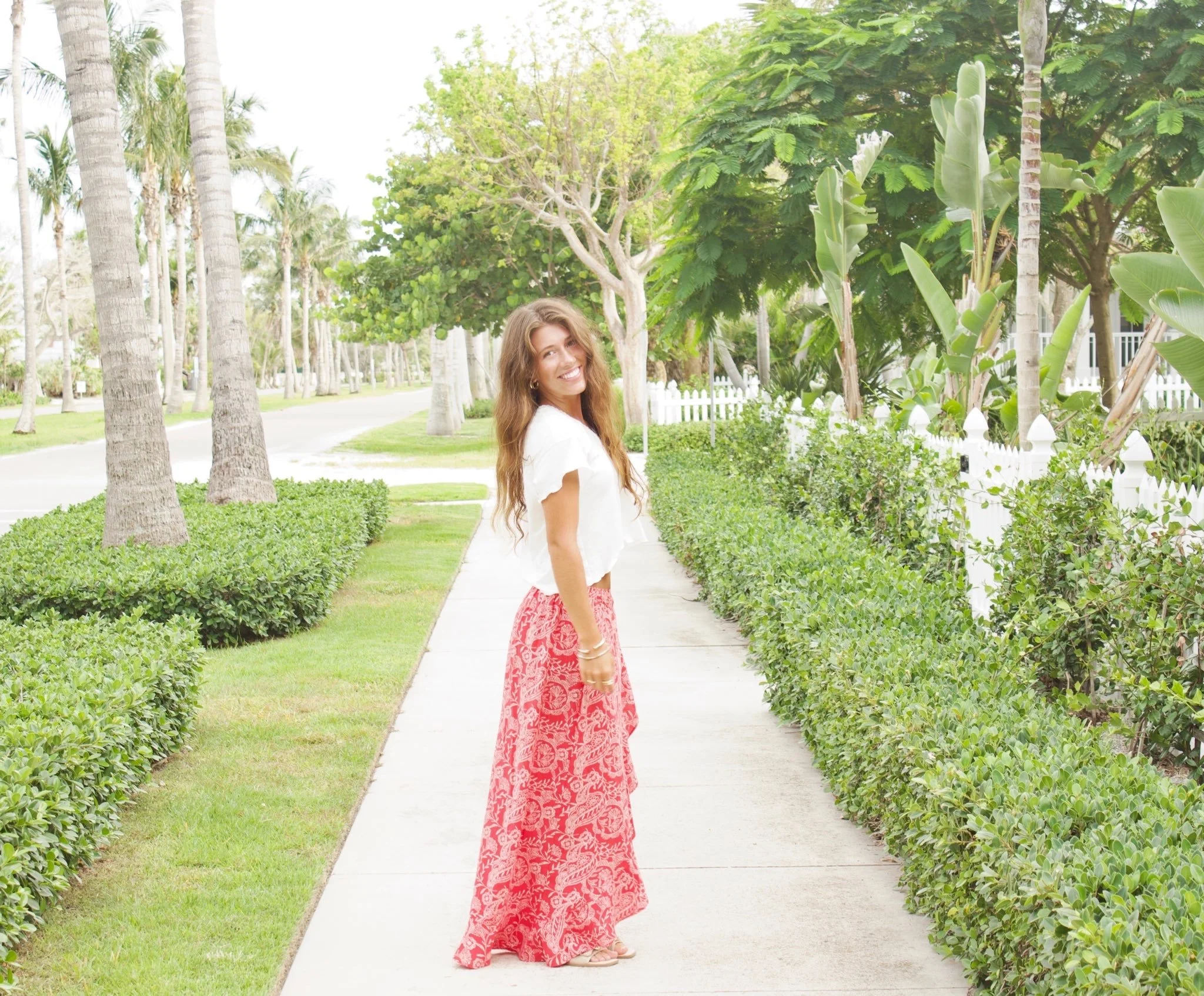 A woman with long brown hair smiling, wearing a white top and red patterned skirt, standing on a sidewalk surrounded by lush green plants and tall palm trees, with a white picket fence in the background.