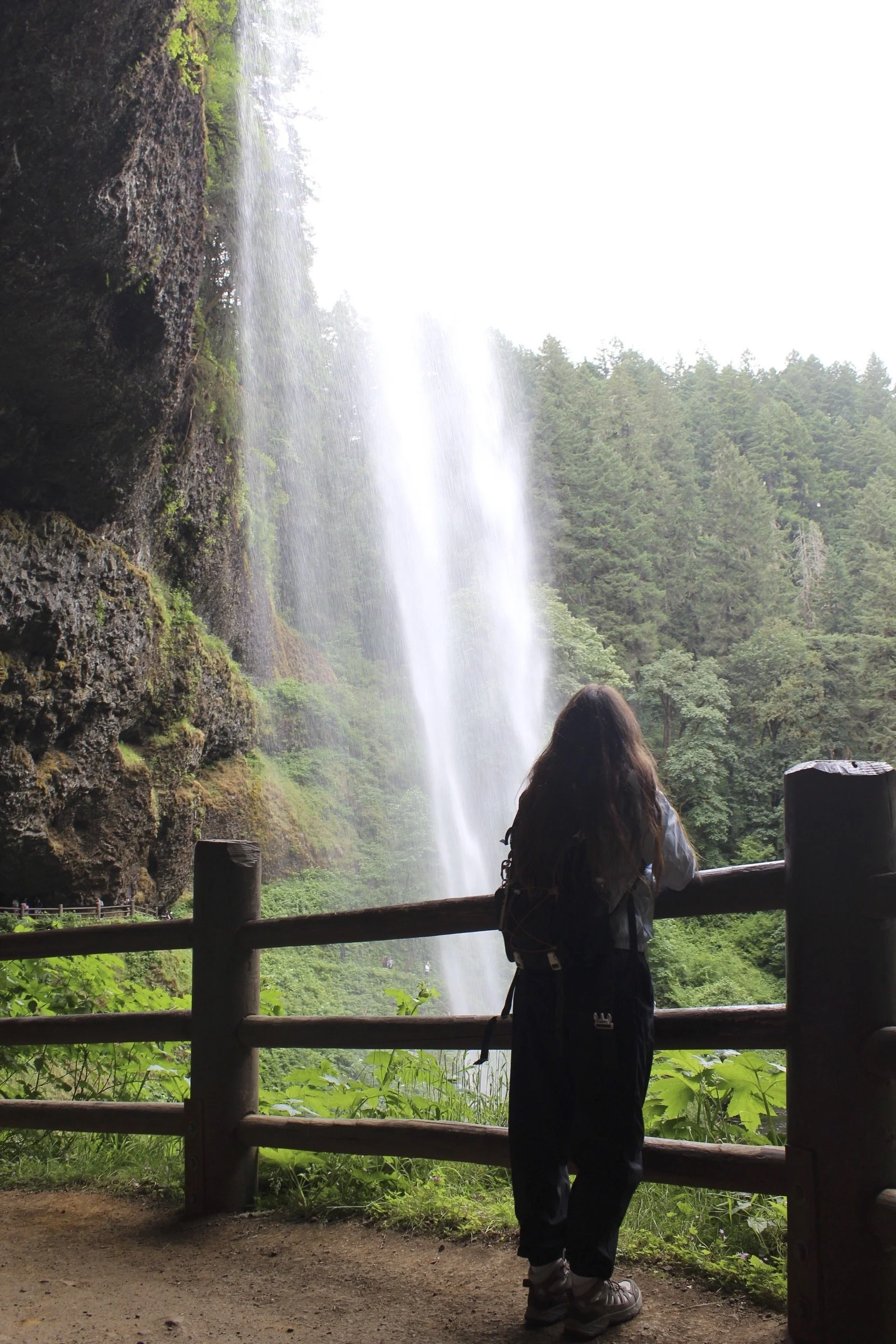A person with long hair, dressed in outdoor clothing, standing behind a wooden railing and looking at a waterfall surrounded by lush green trees.