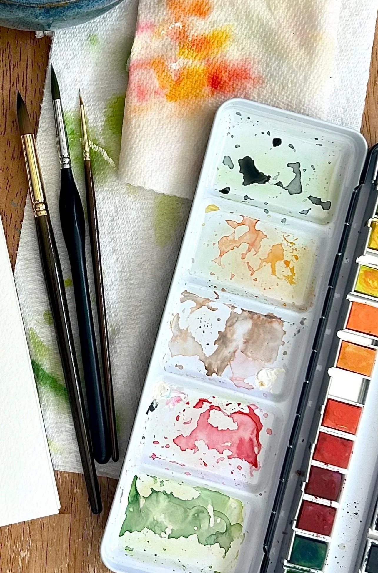 Watercolor paints palette, brushes, and a watercolor paper with colorful paint smudges. The palette has multiple wells with dried watercolor paint, and there are brushes with watercolor paint on the paper towel.