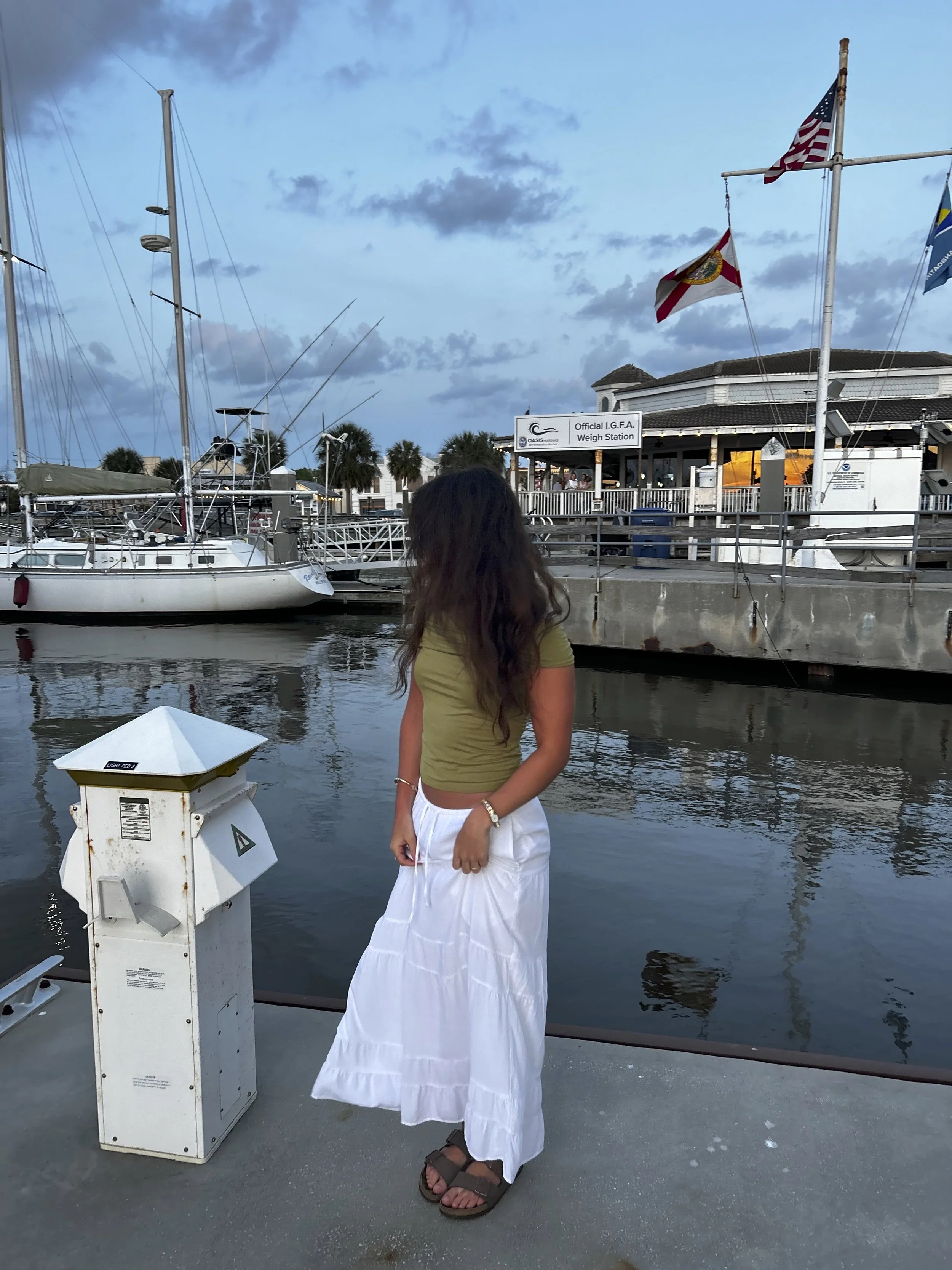 Person with long dark hair wearing a green top and white skirt standing on a dock near a boat, with flags flying on the pier and a building in the background.