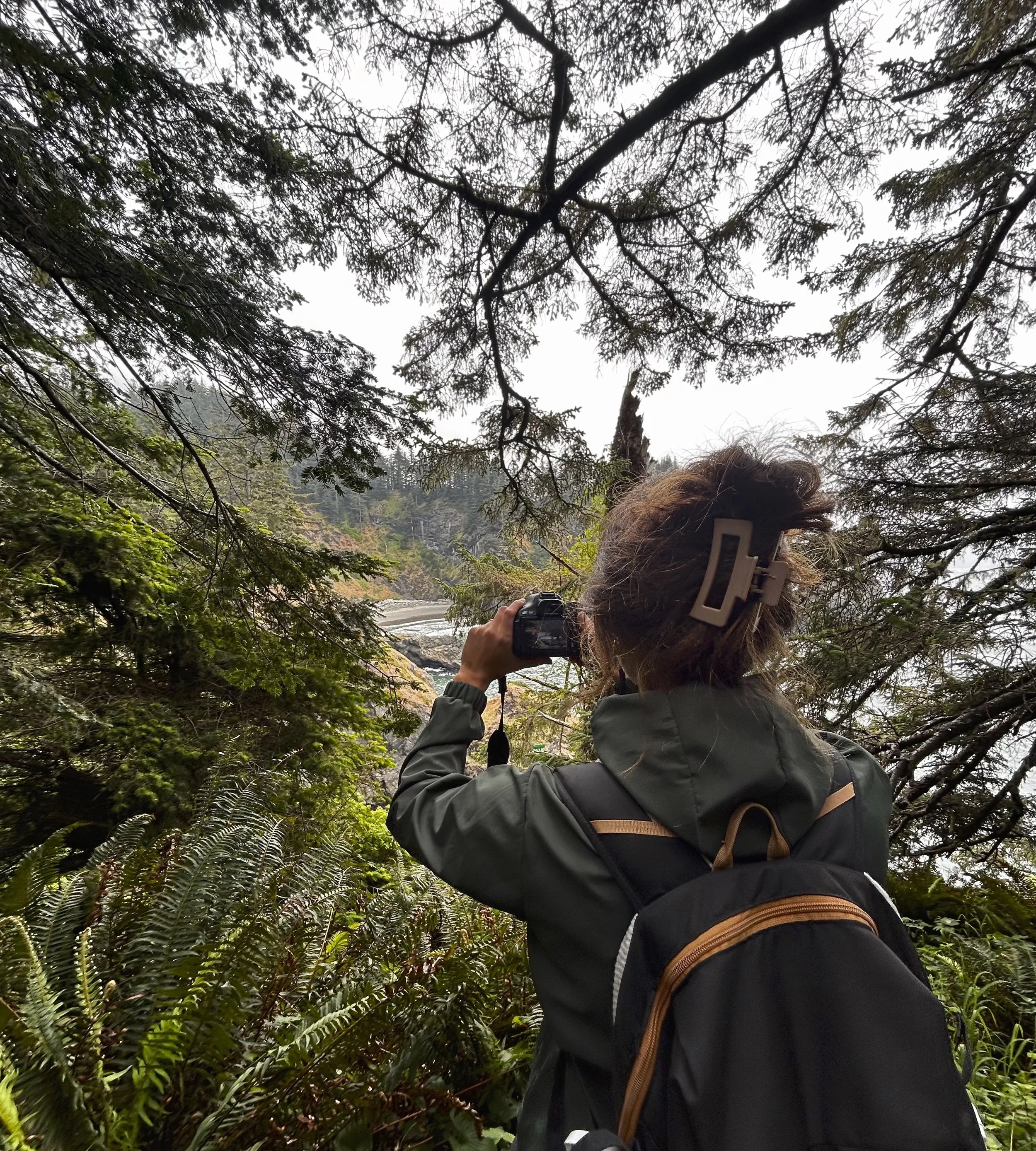Person with brown hair and a dark jacket taking a photograph in a forestedarea, with trees and foliage around and a cloudy sky overhead.