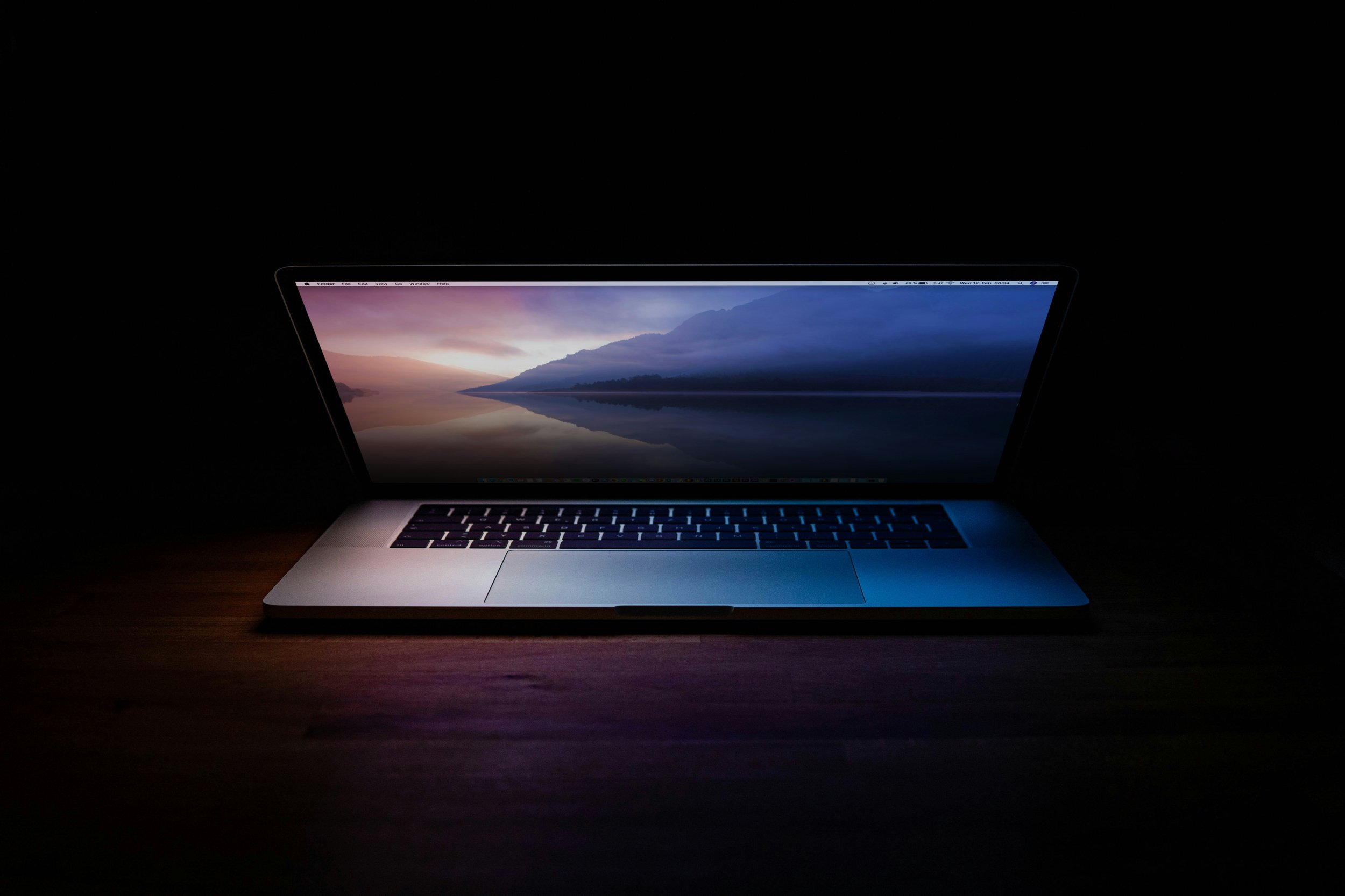 A laptop with a landscape screen displaying mountains and a body of water, on a wooden surface in a dark setting.