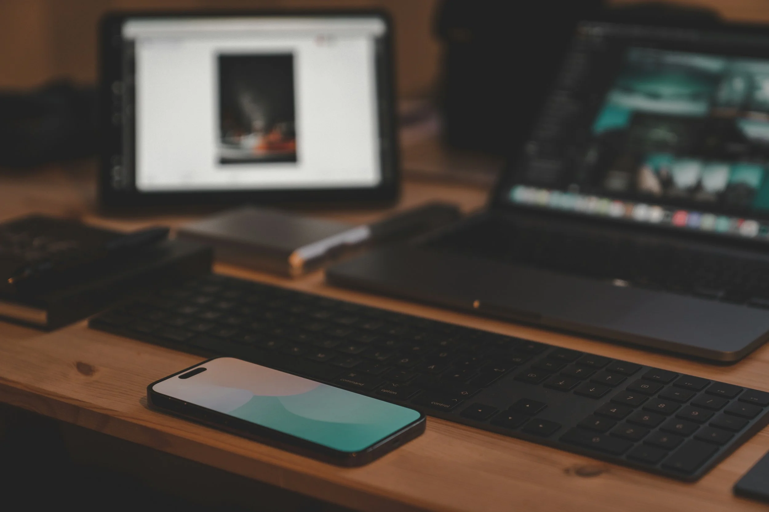 Blurred image of a workspace with a computer monitor, keyboard, smartphone, and multiple electronic devices on a wooden desk.