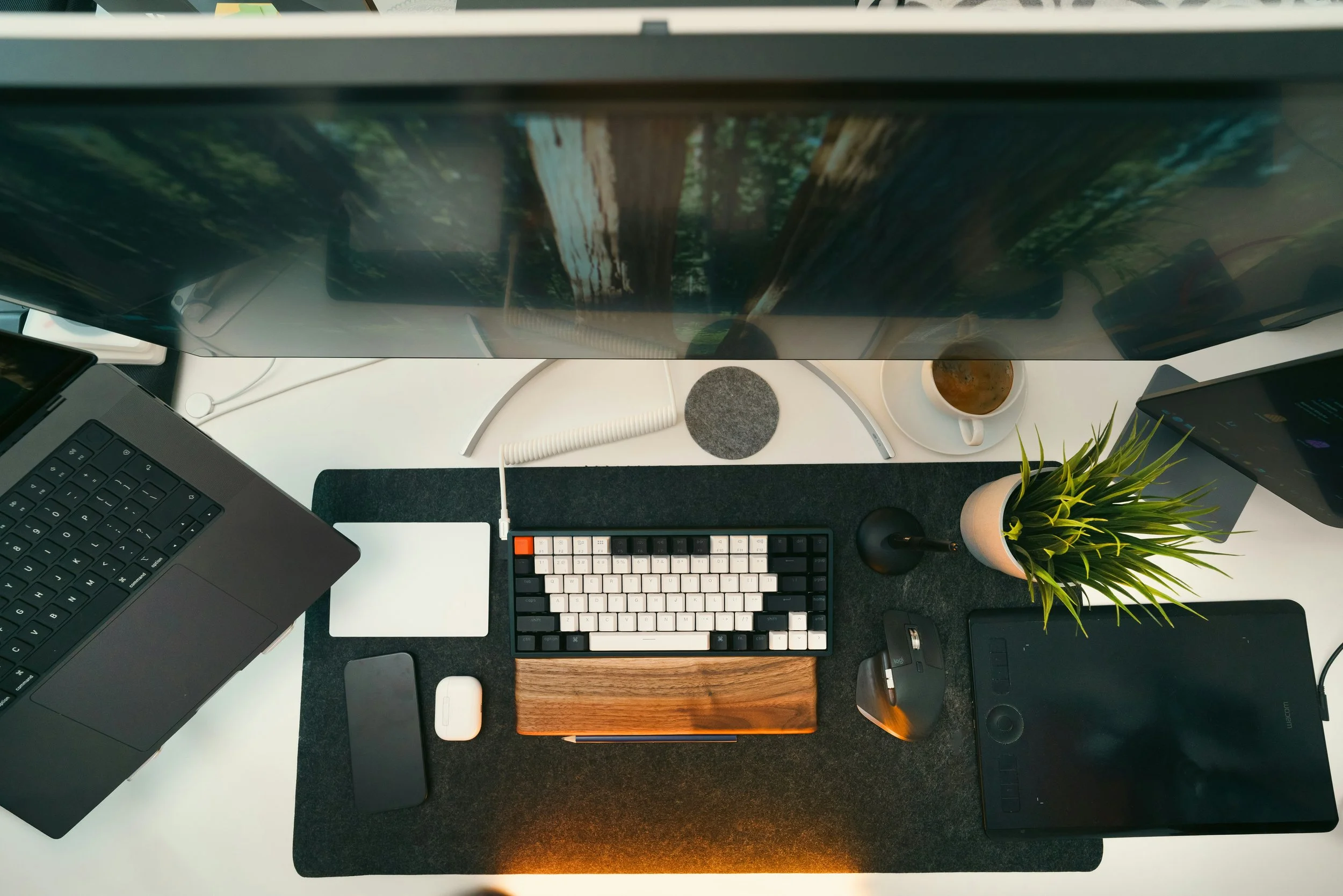 A top-down view of a workspace with a computer monitor, a laptop, a wireless keyboard, a white wireless mouse, a small potted plant, a coffee cup, and various electronic accessories on a white desk with a black desk mat.