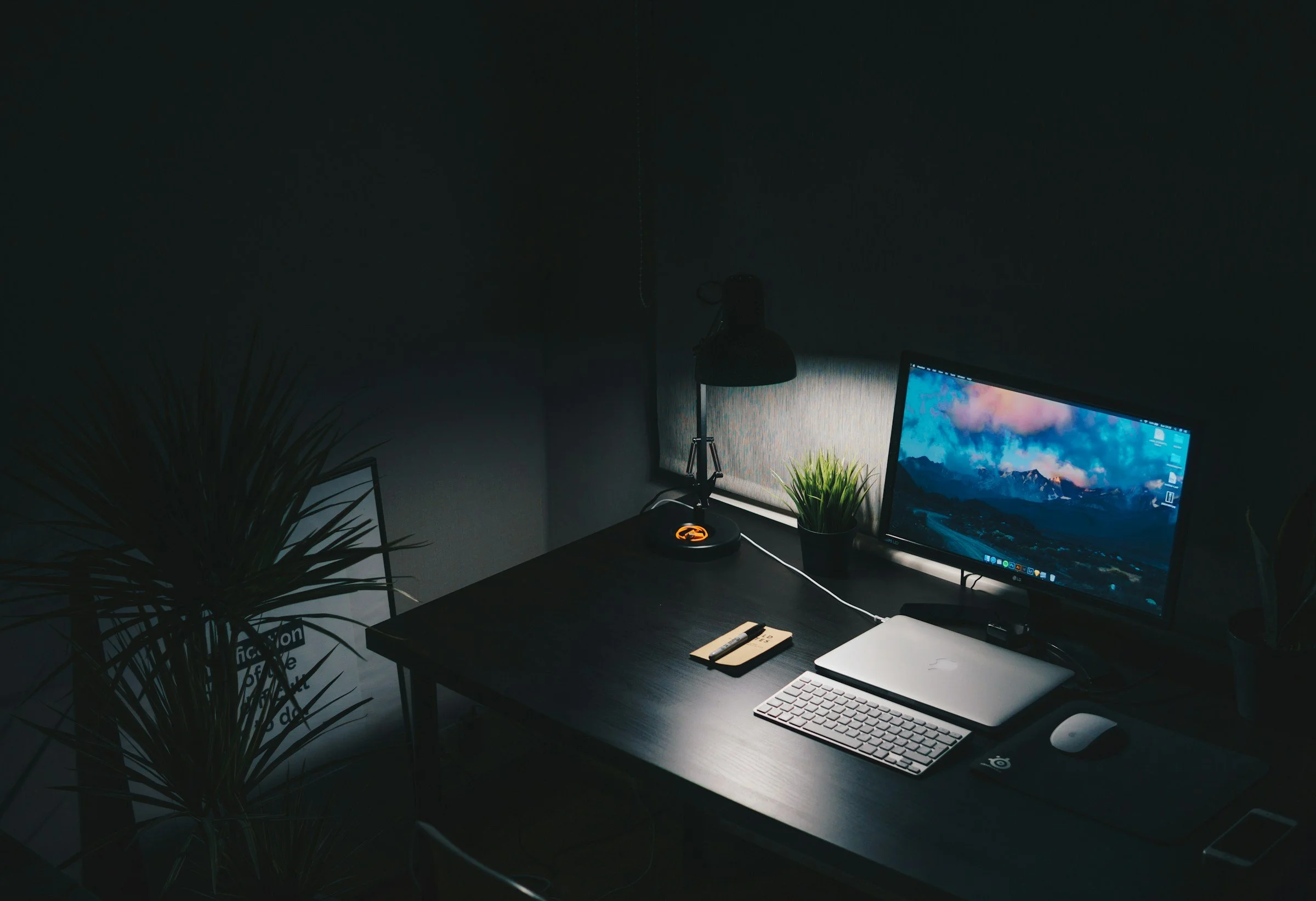 A dark home office setup with a computer monitor displaying a mountain landscape, a keyboard, a mouse, a notepad with a pen, a desk lamp, a small potted plant, and some wall decorations, with natural light coming from a window.
