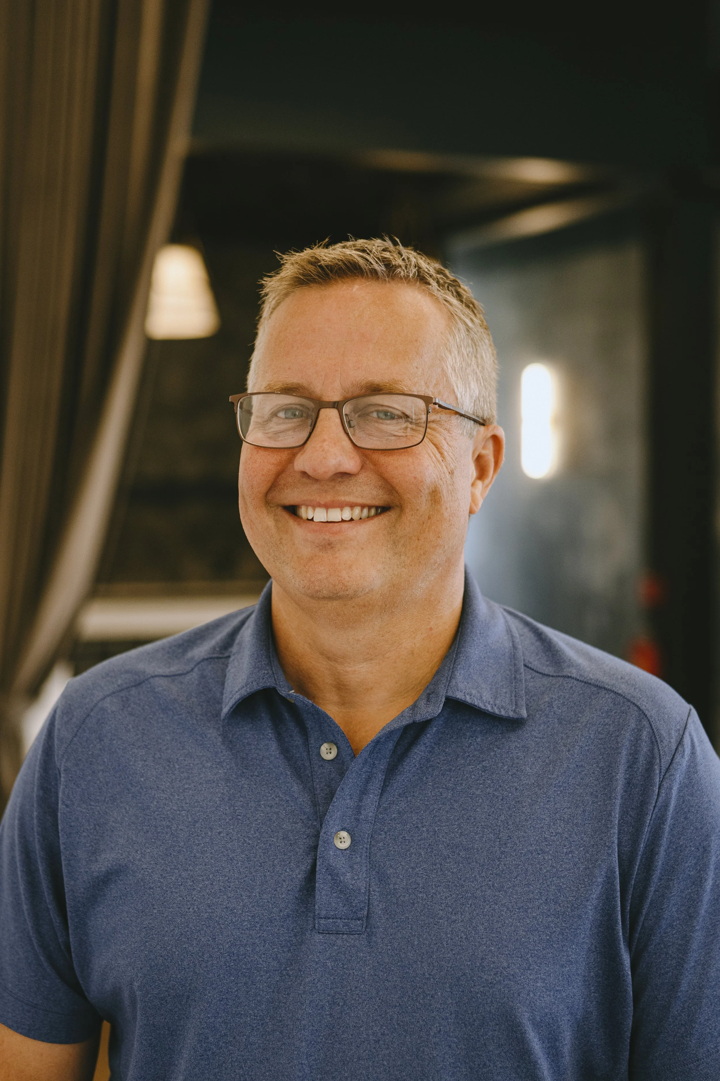 A smiling man with glasses and short blonde hair wearing a dark blue polo shirt, indoors with dark and warm lighting.