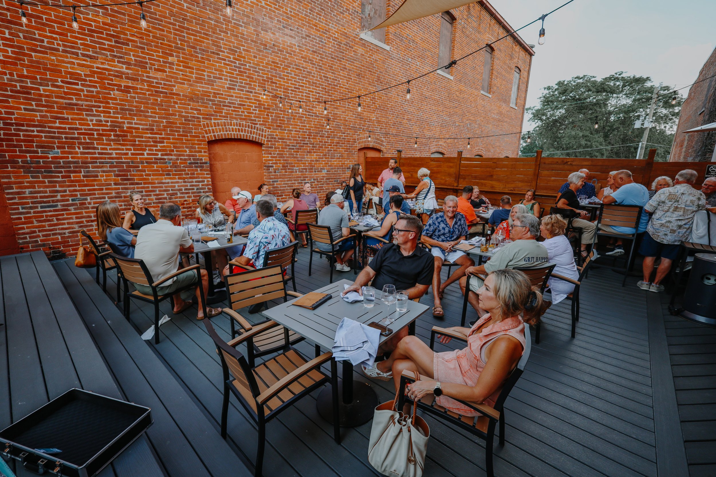 People dining outdoors on a patio with brick walls, wooden fencing, tables, and chairs, with string lights overhead.