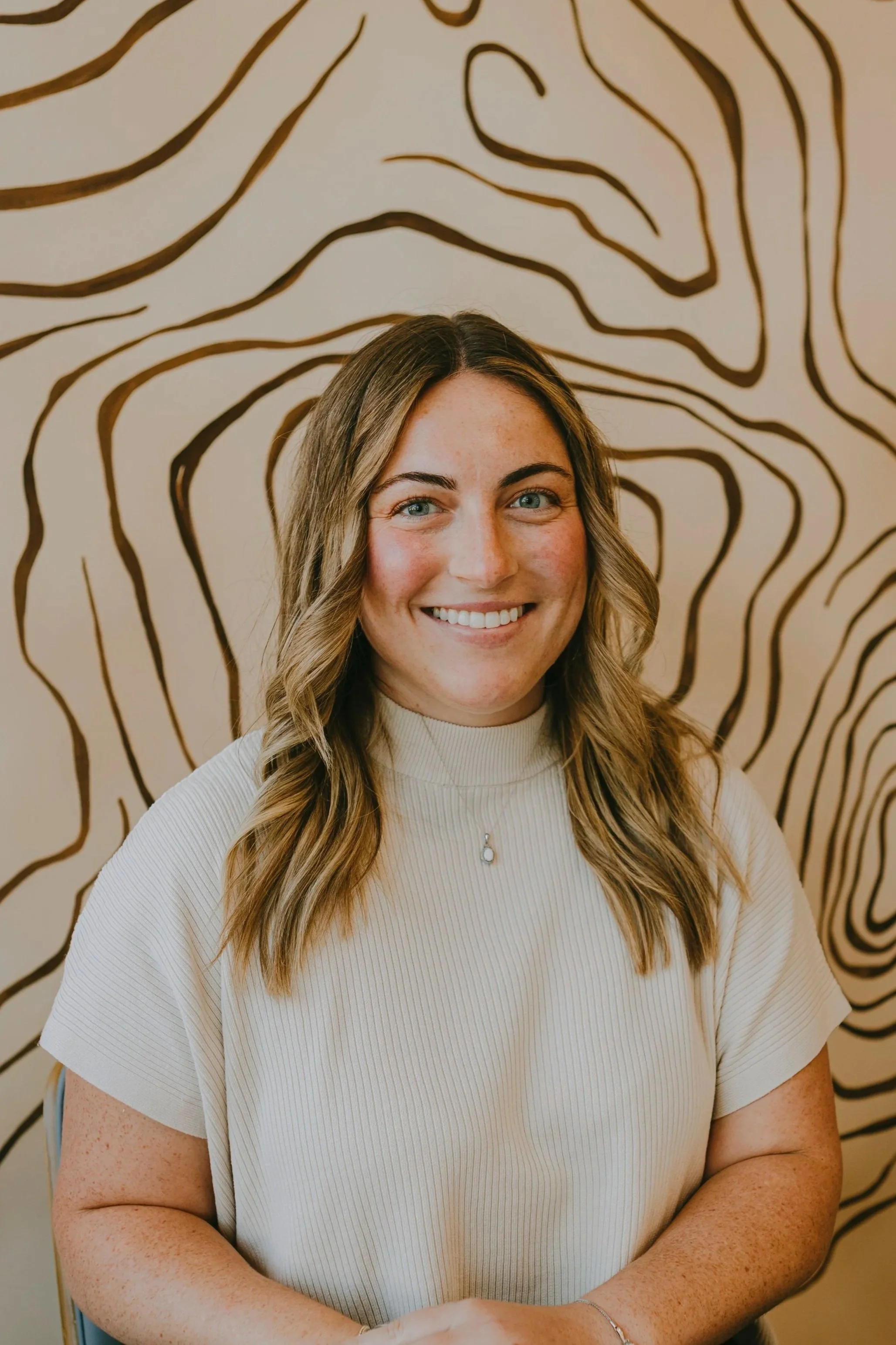 A smiling woman with long, wavy blonde hair and blue eyes, wearing a cream-colored ribbed top, sitting in front of a wall with black and brown abstract line art.