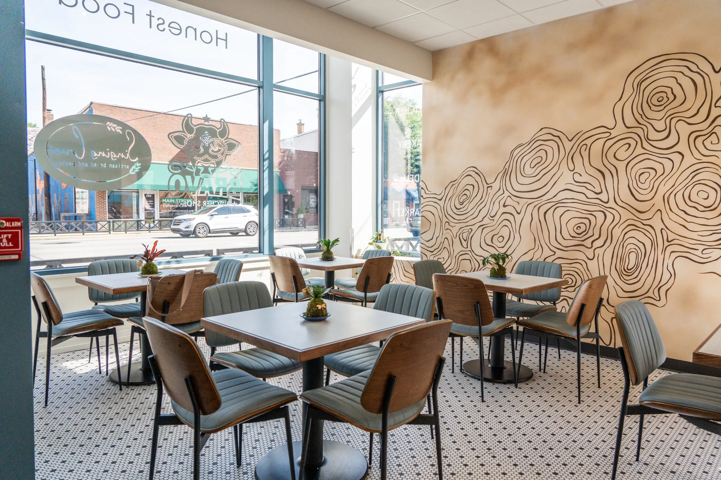 Inside a cafe with wooden chairs and tables decorated with small plants, large windows letting in natural light, and a wall with a topographic map design. Outside the window, a street with parked cars and brick buildings is visible.
