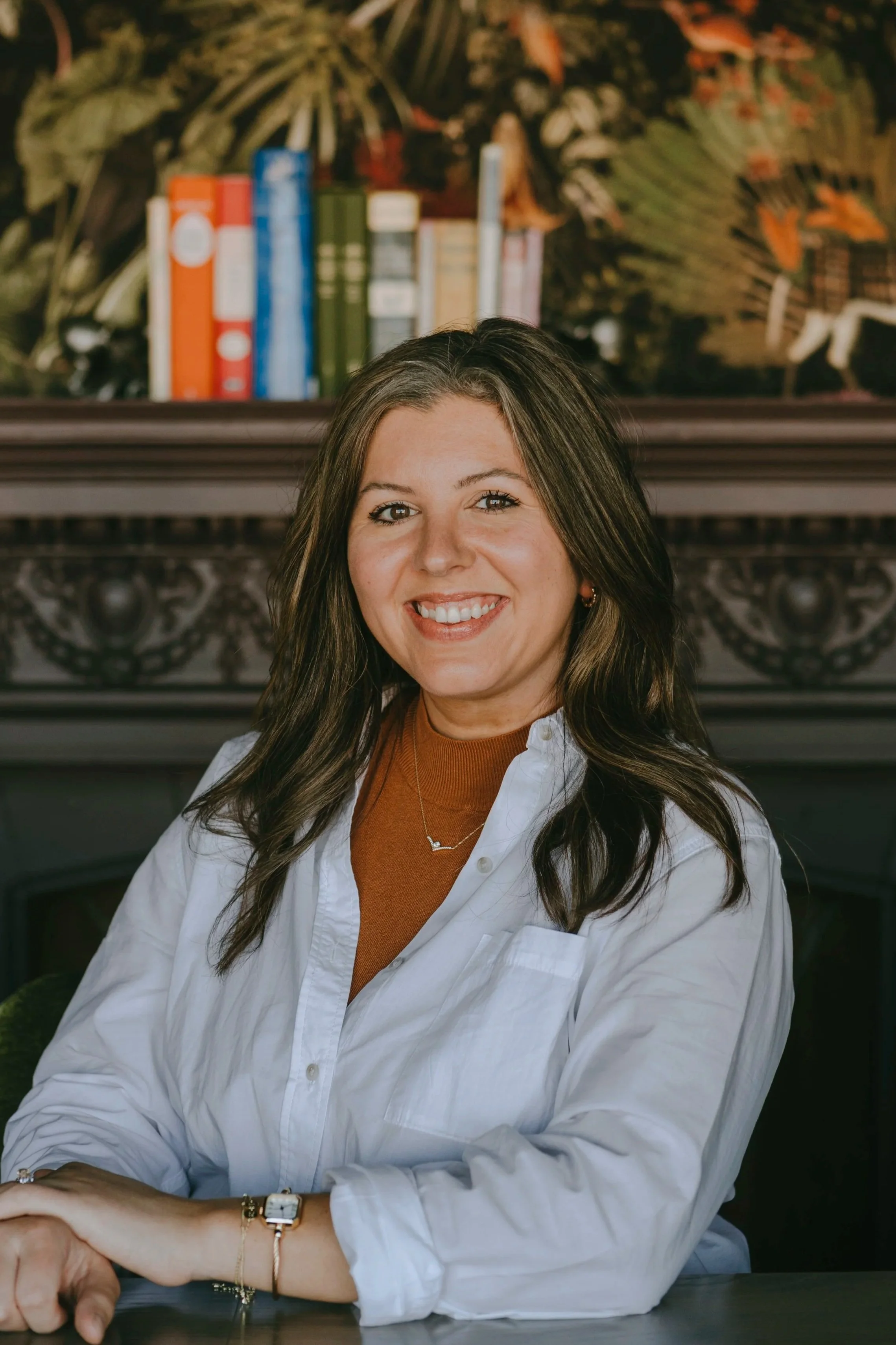 A woman with long dark hair smiling while sitting at a table, wearing a white shirt and a brown top, with a background of a bookshelf and decorative woodwork.