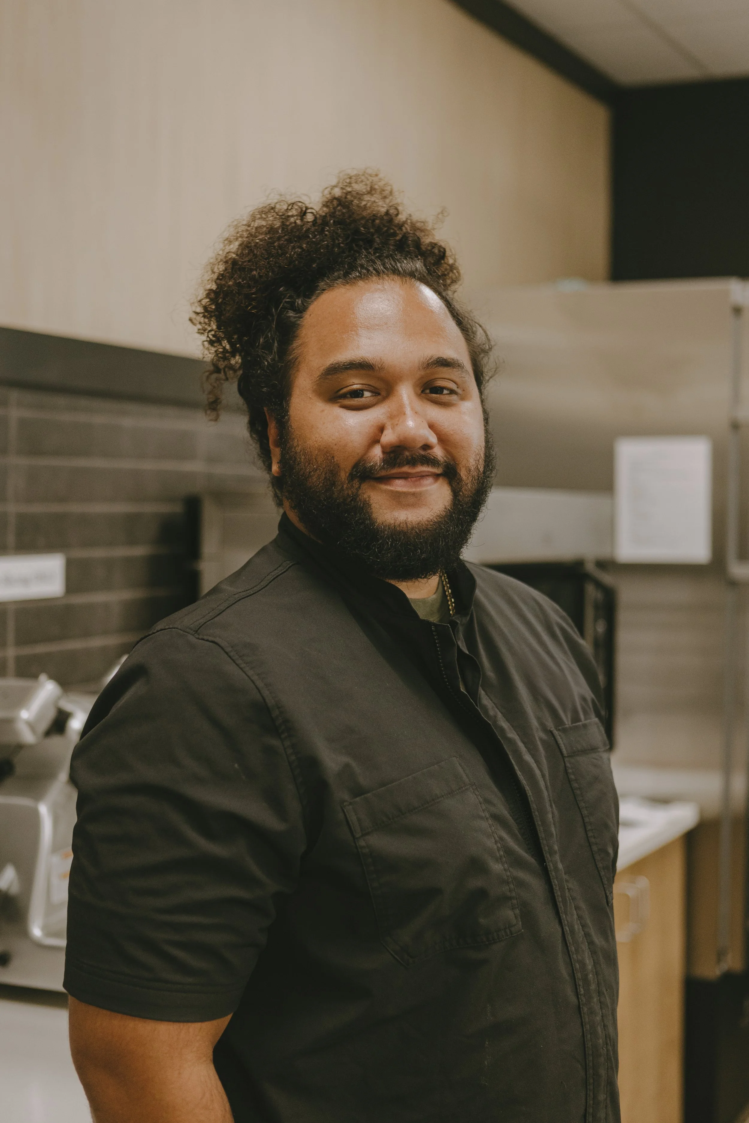 A man with curly hair, a beard, and dark clothing standing in a kitchen with cabinets and appliances in the background.
