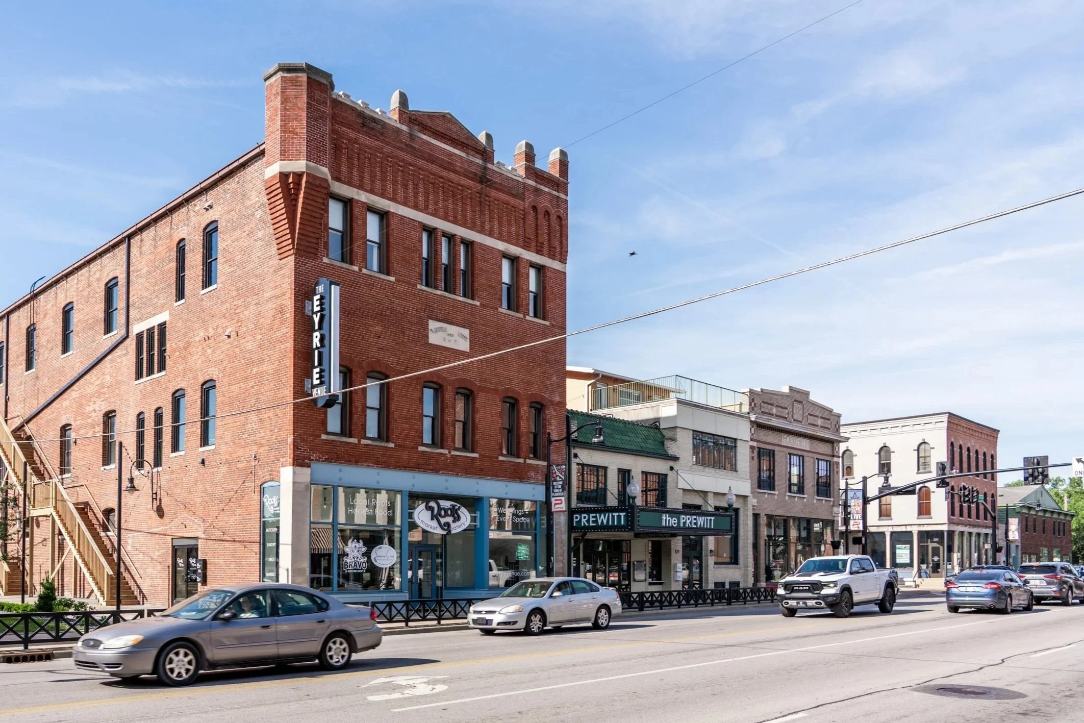 Street view of a historic red brick building with various storefronts, cars parked along the street, and a clear blue sky overhead.