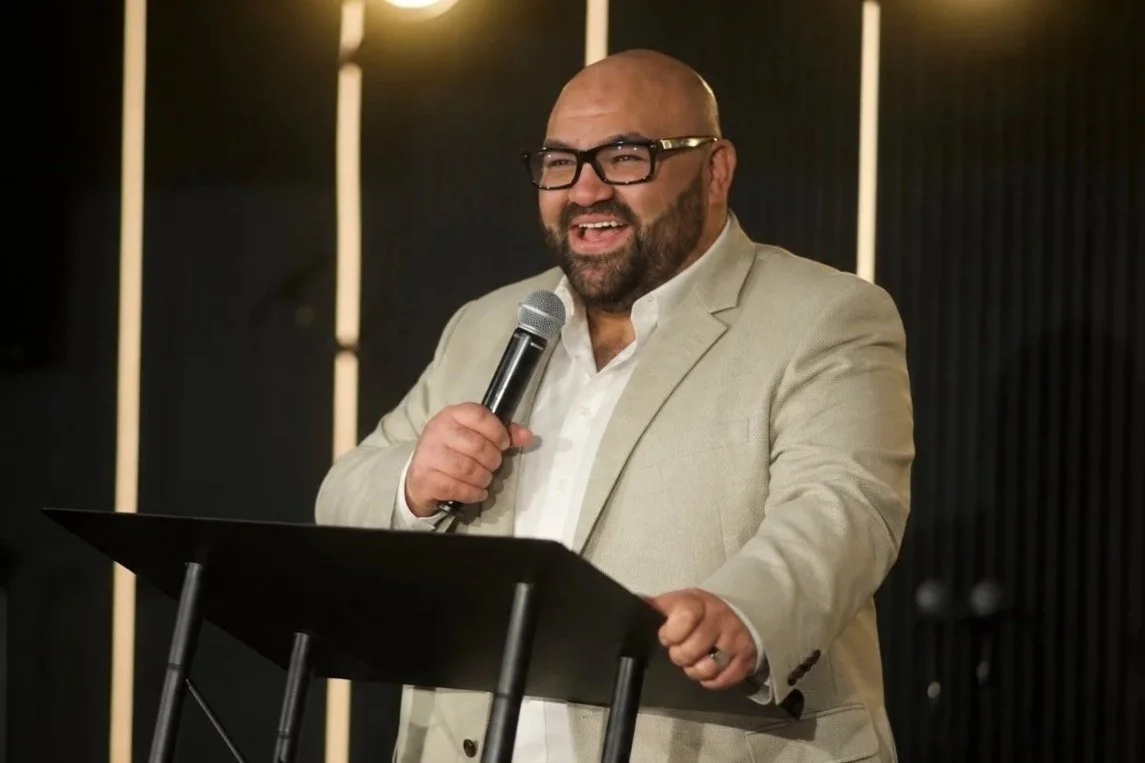 A man with a beard and glasses, wearing a beige suit, speaking into a microphone at a podium, smiling.