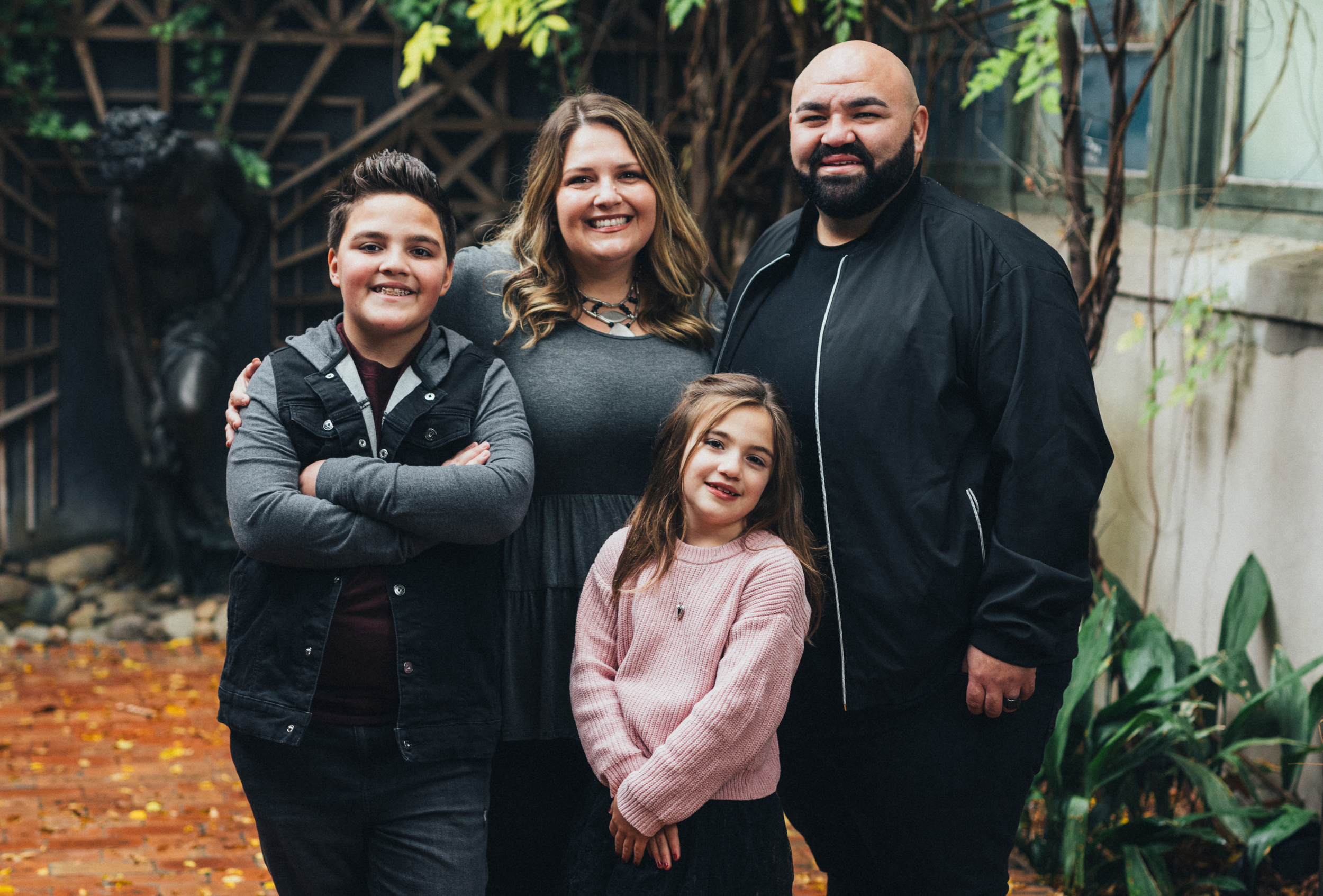 Family of five standing outdoors, smiling at the camera. They are surrounded by greenery with a brick path and trees in the background. The group includes a woman, a man, two girls, and a boy.