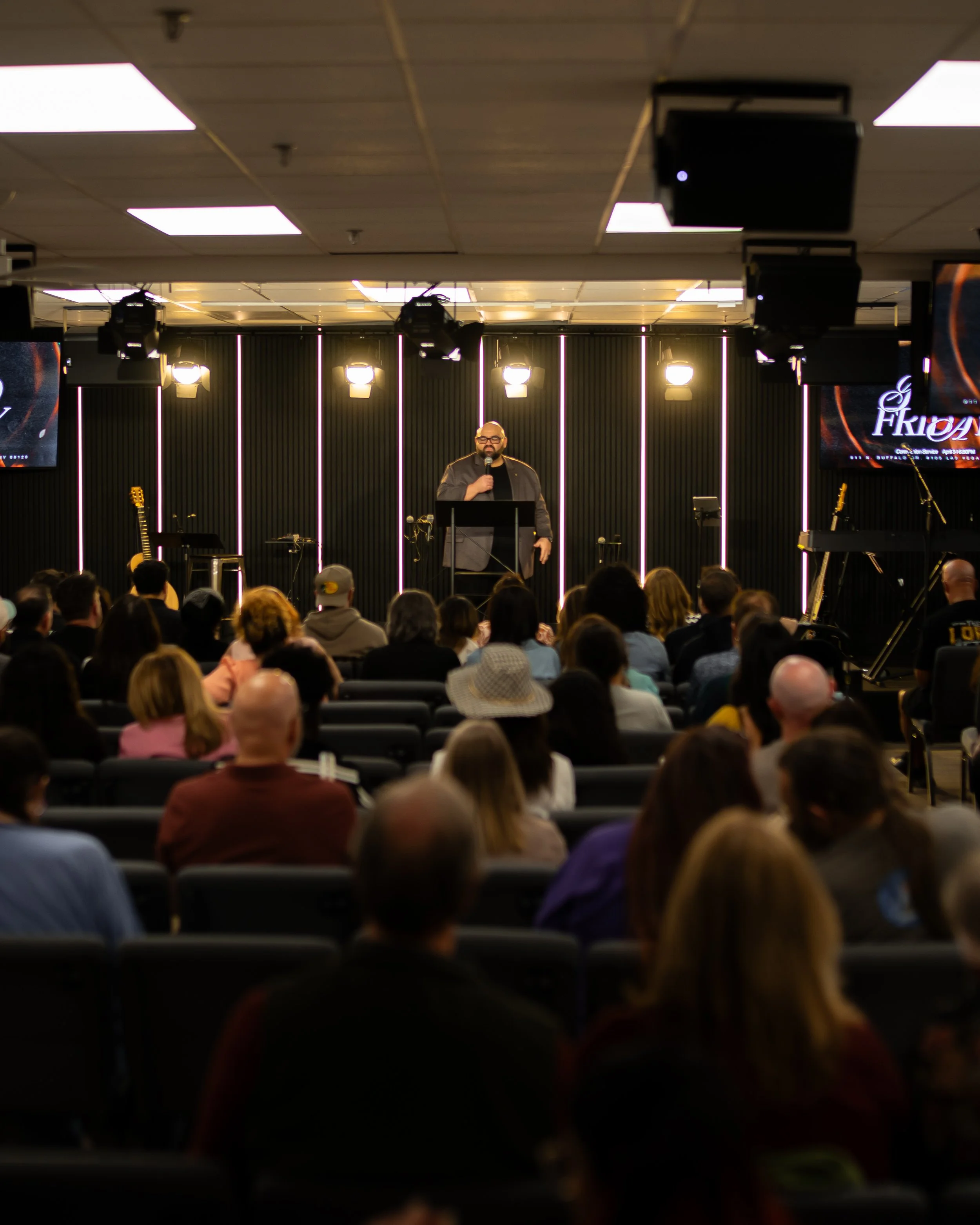 A man standing on a stage with a microphone in hand, addressing an audience in a conference room with black walls and vertical light strips, with guitars and sound equipment on stage.