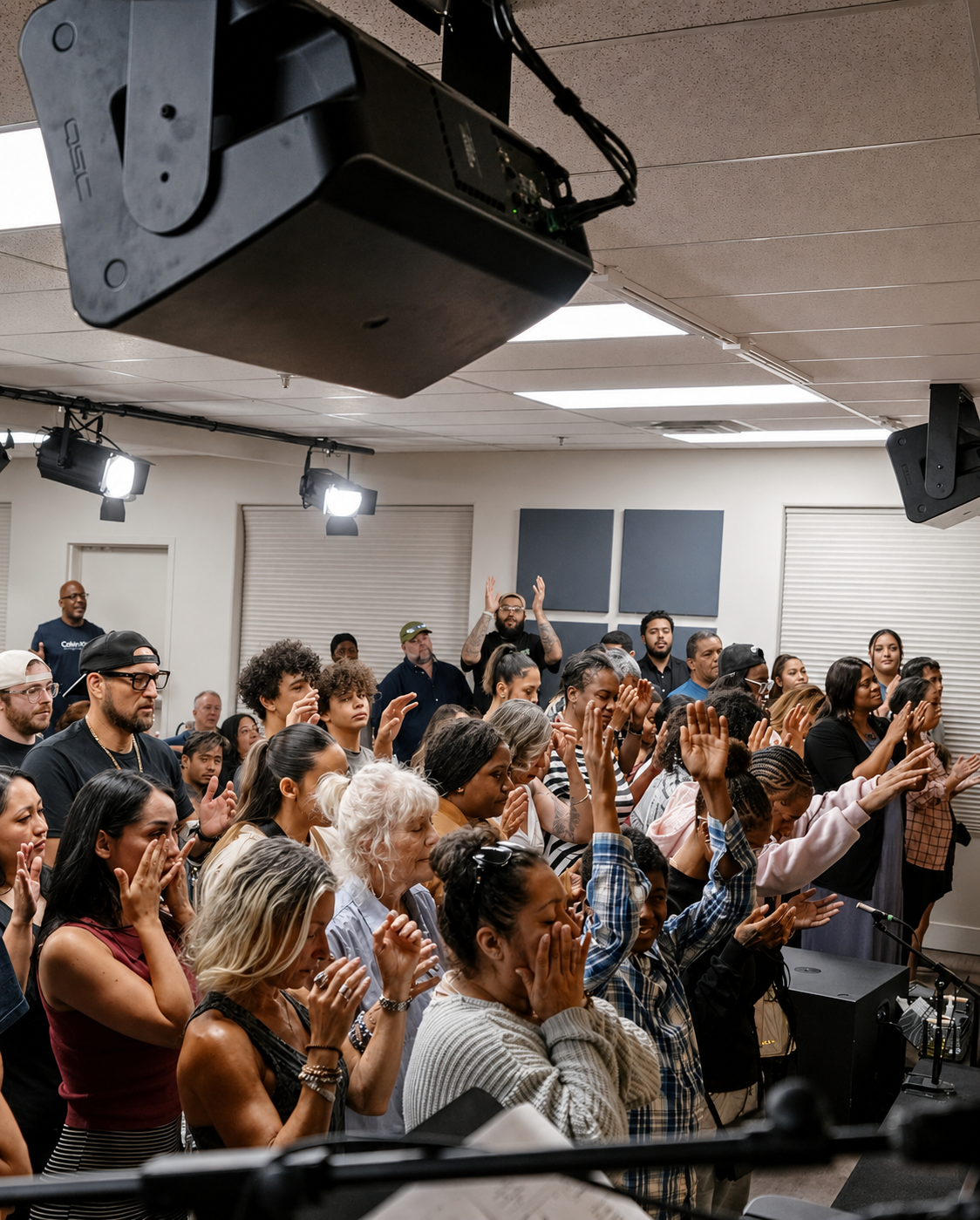A diverse group of people gathered in a room, standing with many raising their hands and some praying or reflecting, with a few visible on stage and bright lighting overhead.