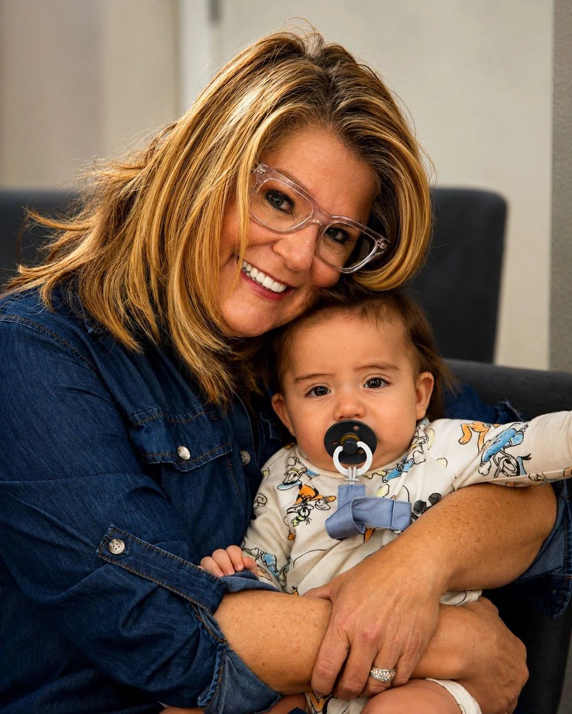 A woman with blonde hair, glasses, and a denim shirt holding a young child with a pacifier in their mouth. The woman is smiling warmly, and the child is looking at the camera with a neutral expression.