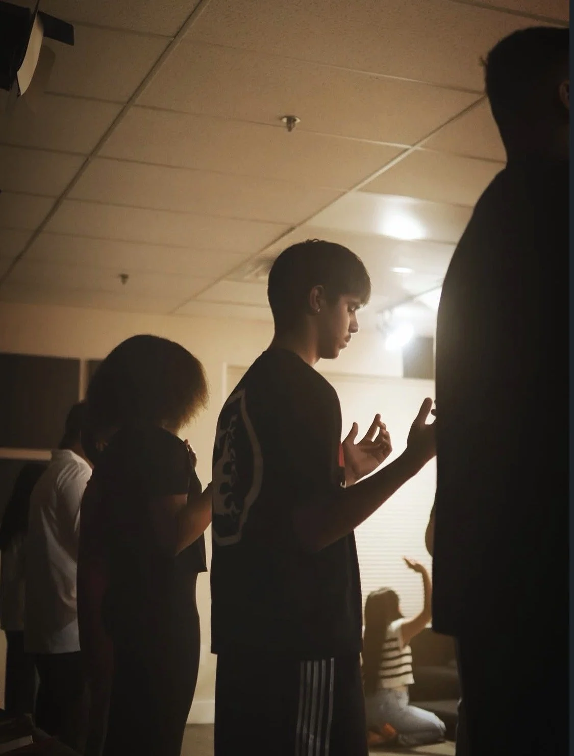 People standing in prayer, with some kneeling and others with hands raised or held in meditative gestures, inside a room with a low ceiling and soft lighting.
