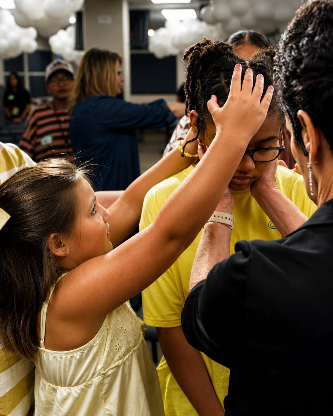 Young girl places her hand on a teenager's head as part of a prayer circle in an indoor setting with clouds hanging from the ceiling.