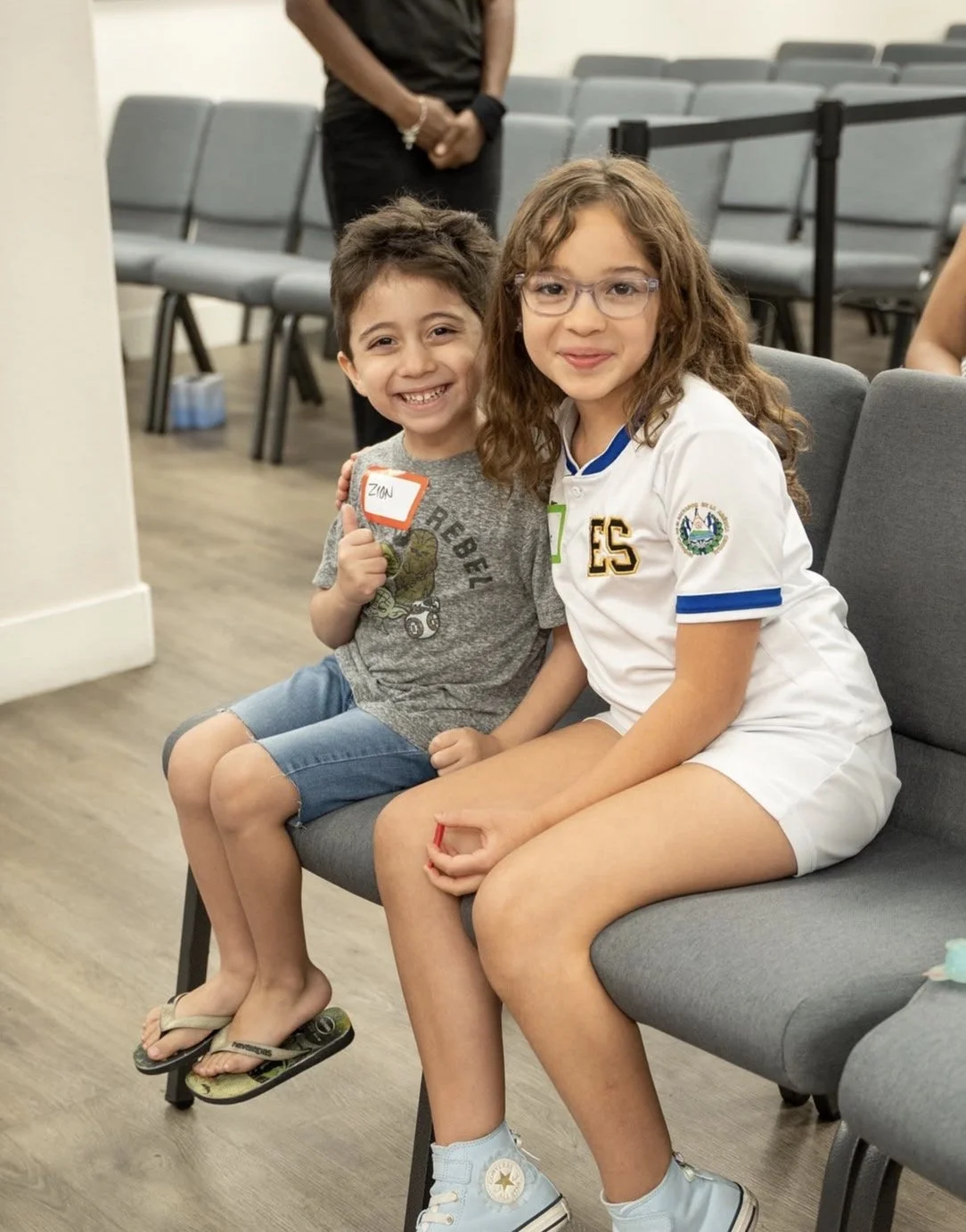 Two children, a boy and a girl, sitting on a gray bench in what appears to be a waiting area or auditorium. The boy, holding a name tag that says 'Zion,' is wearing a gray T-shirt, blue shorts, and flip-flops. The girl, wearing glasses and a white sports jersey, is dressed in white shorts and light blue Converse sneakers. They are smiling and sitting close together, with the girl’s arm around the boy.