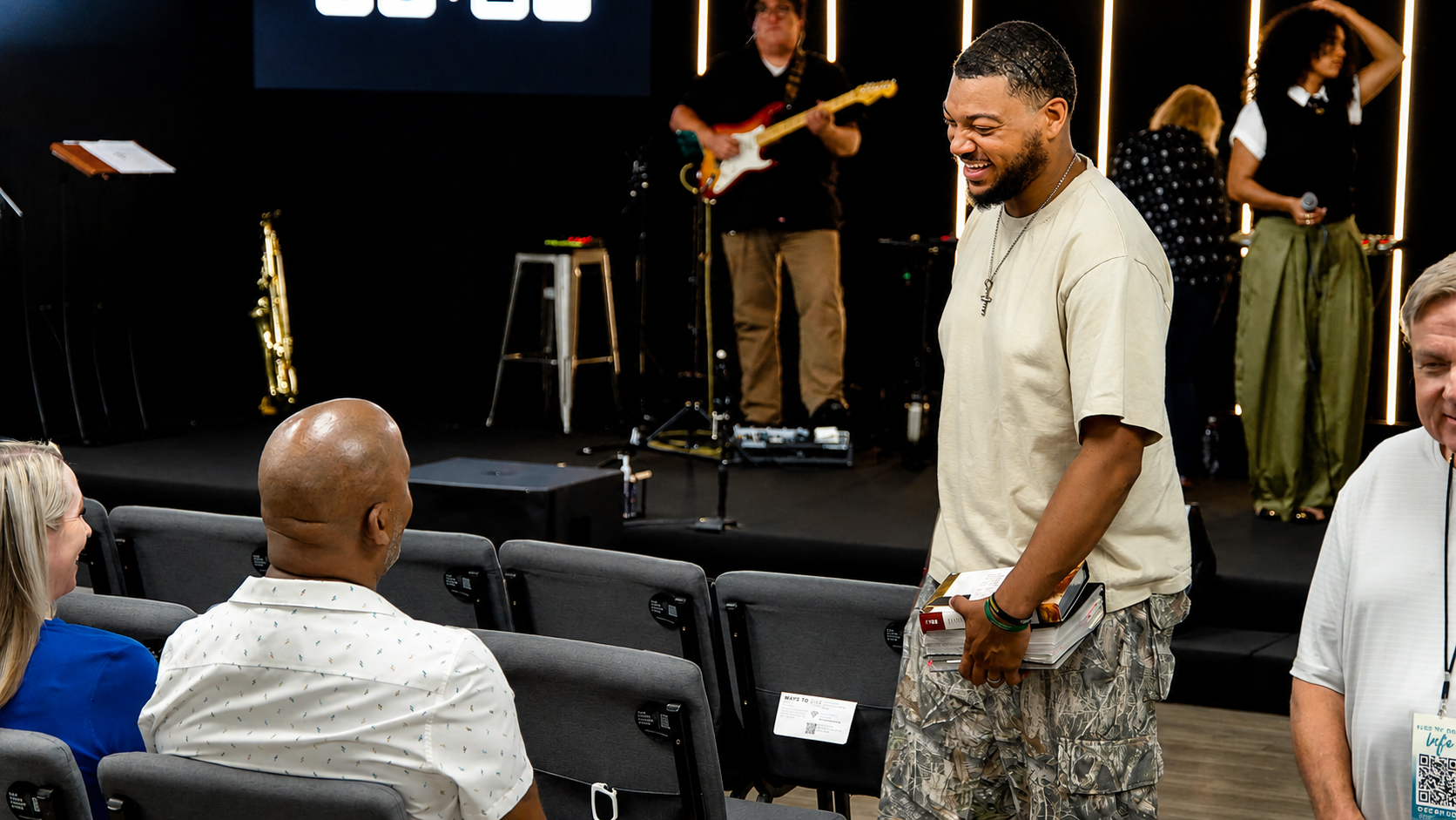 A man with a beard, wearing a beige T-shirt and camo shorts, stands smiling and talking to a seated woman and a man with a shaved head, in an indoor event space with a stage. In the background, there are musicians playing instruments, including a guitarist and a woman with curly hair holding a microphone. The stage has vertical light strips.
