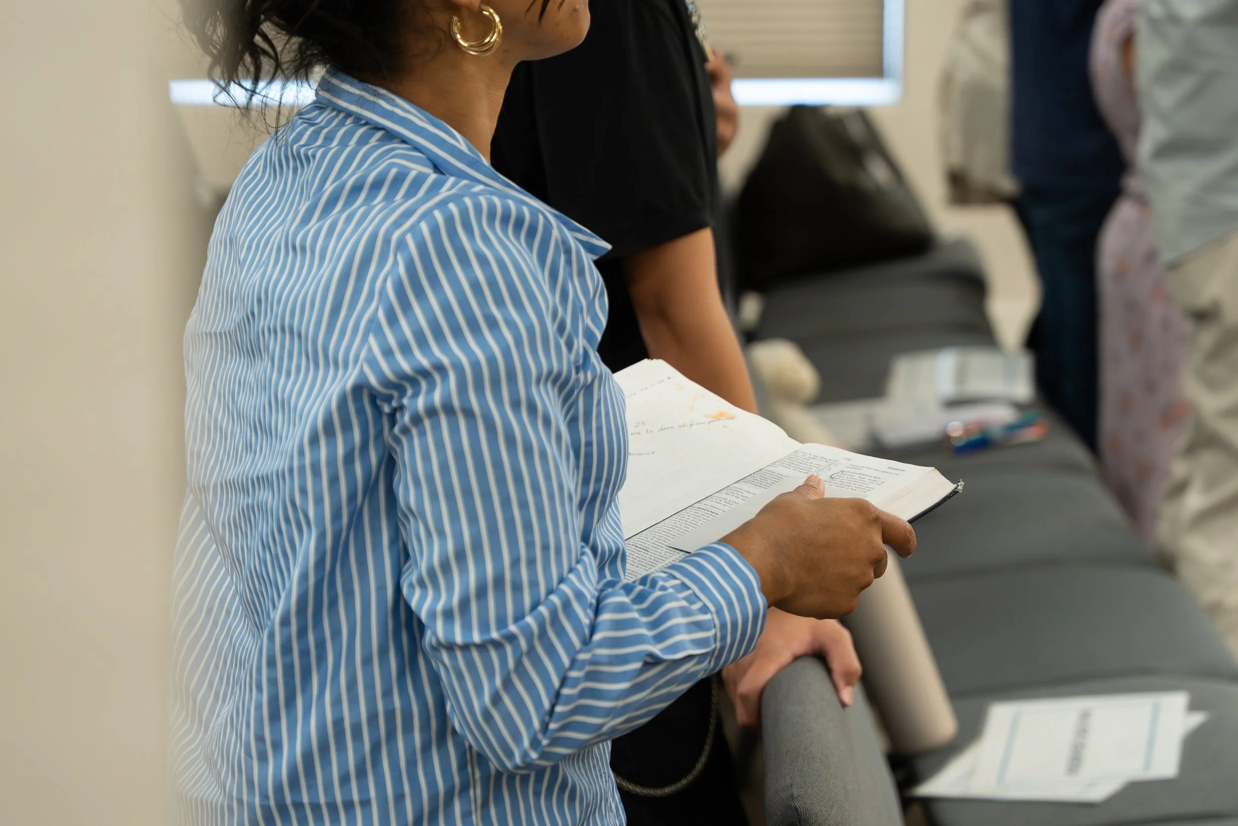 A person wearing a blue and white striped shirt is reading a book, with other people standing nearby in a room with chairs.