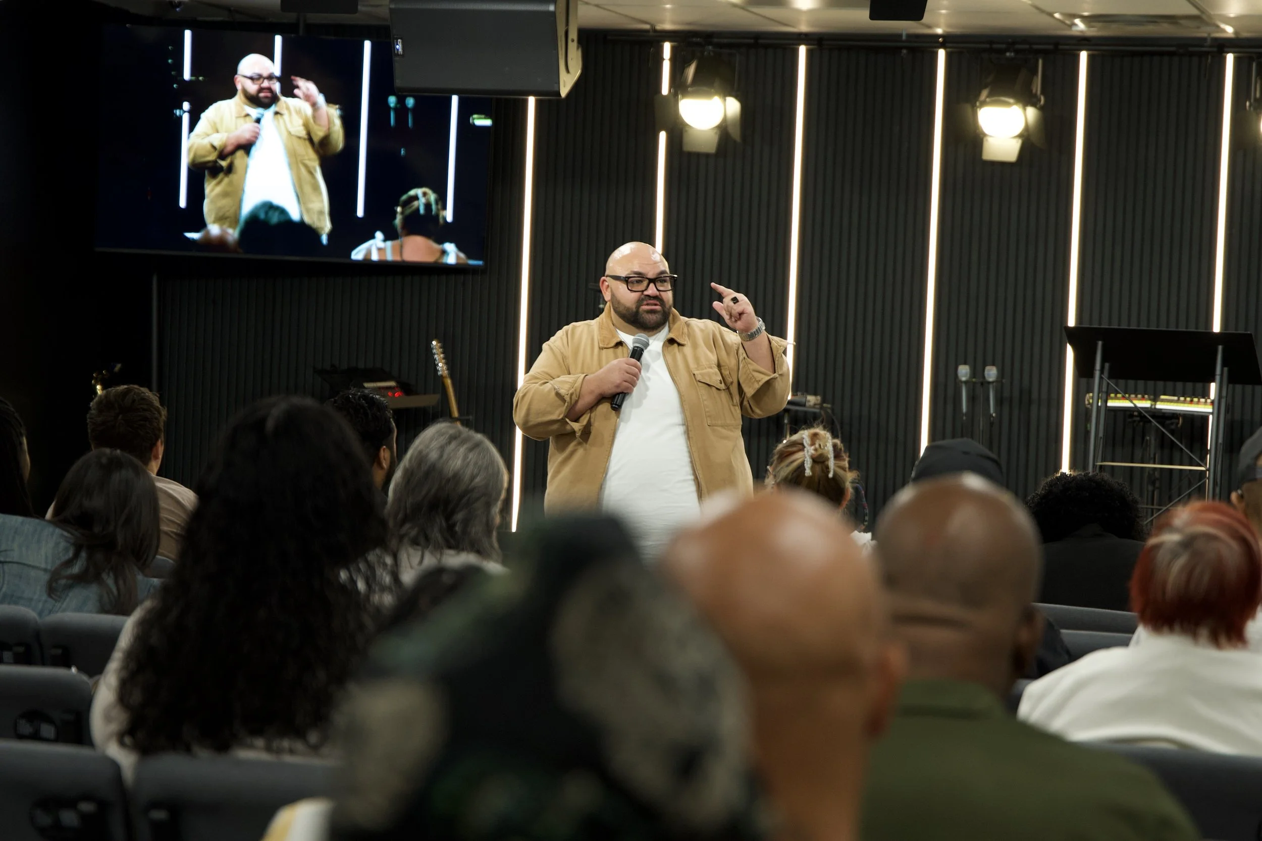 A man with glasses and a beard, dressed in a tan jacket over a white shirt, is speaking into a microphone while gesturing with his right hand during a presentation in a room filled with people. There is a large screen behind him displaying the same man, and the room is decorated with vertical lights and stage lighting.