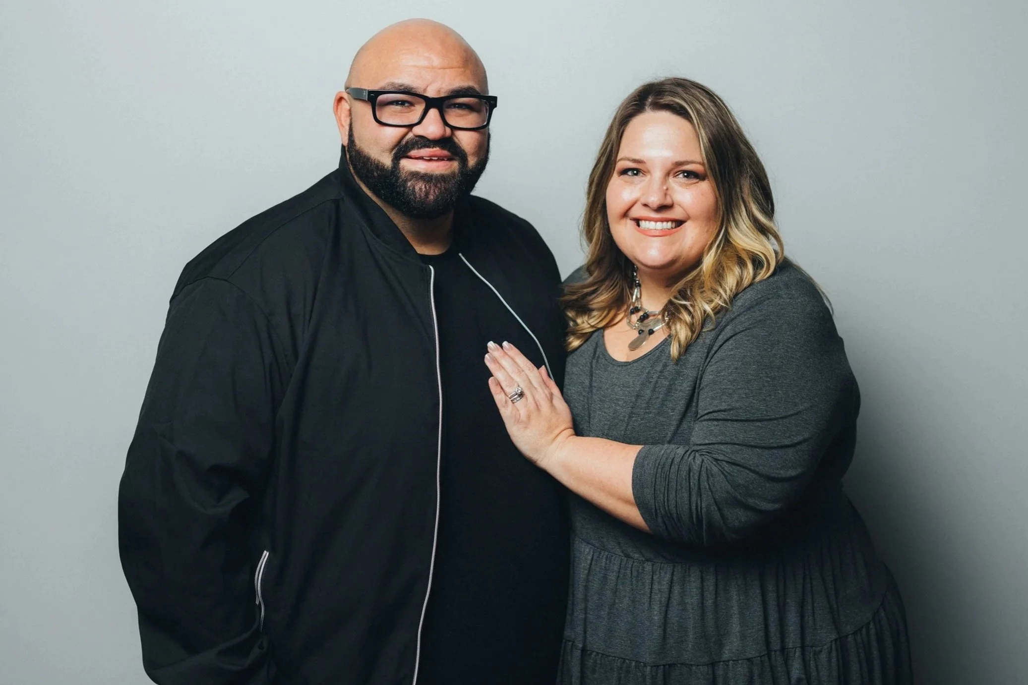 A man with glasses and a beard standing next to a woman with wavy blonde hair, both smiling and touching hands, against a plain light gray background.