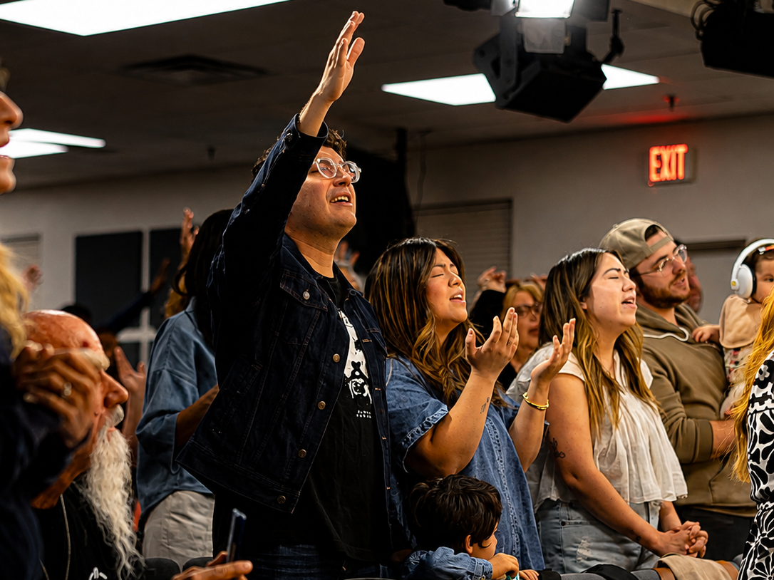 People in a crowd with eyes closed and hands raised, engaged in a spiritual or worship experience inside a church or auditorium.