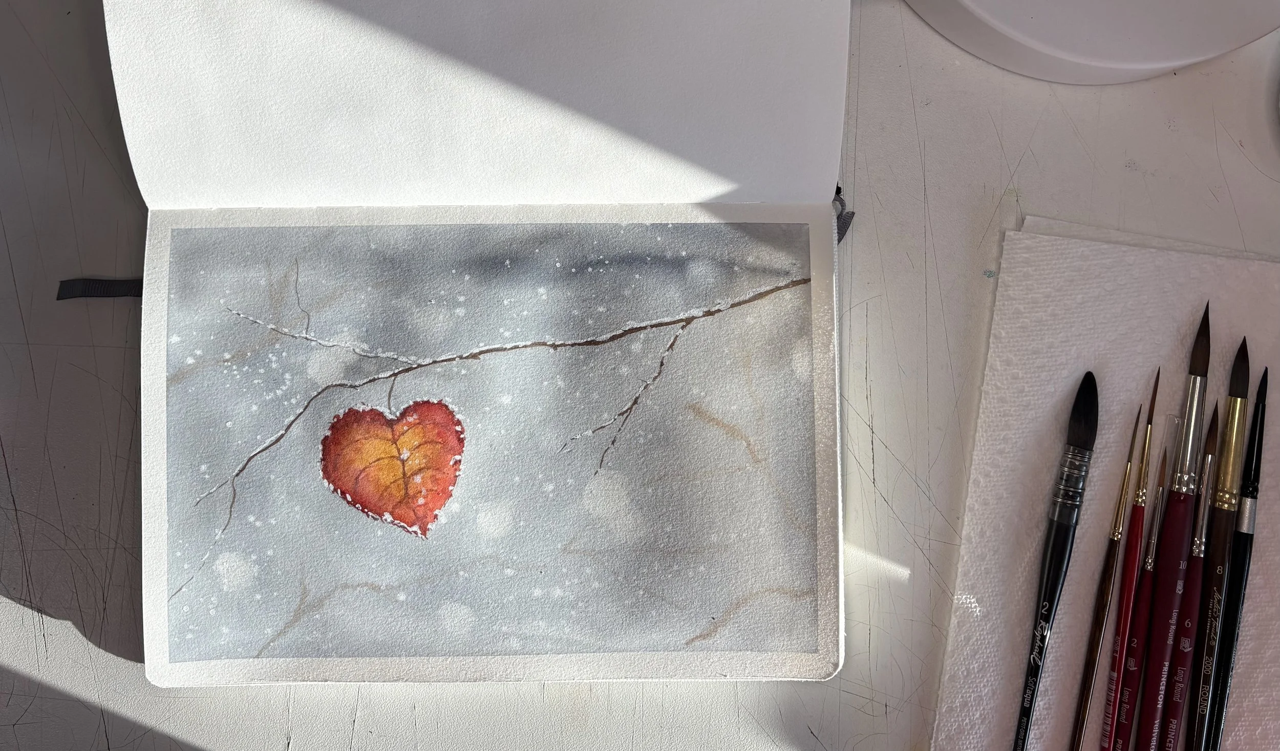 A watercolor painting of a red-orange heart-shaped leaf on a light gray background with cracks and white specks; art supplies including paintbrushes are nearby. This shows my work station as I paint in my Etchr journal.