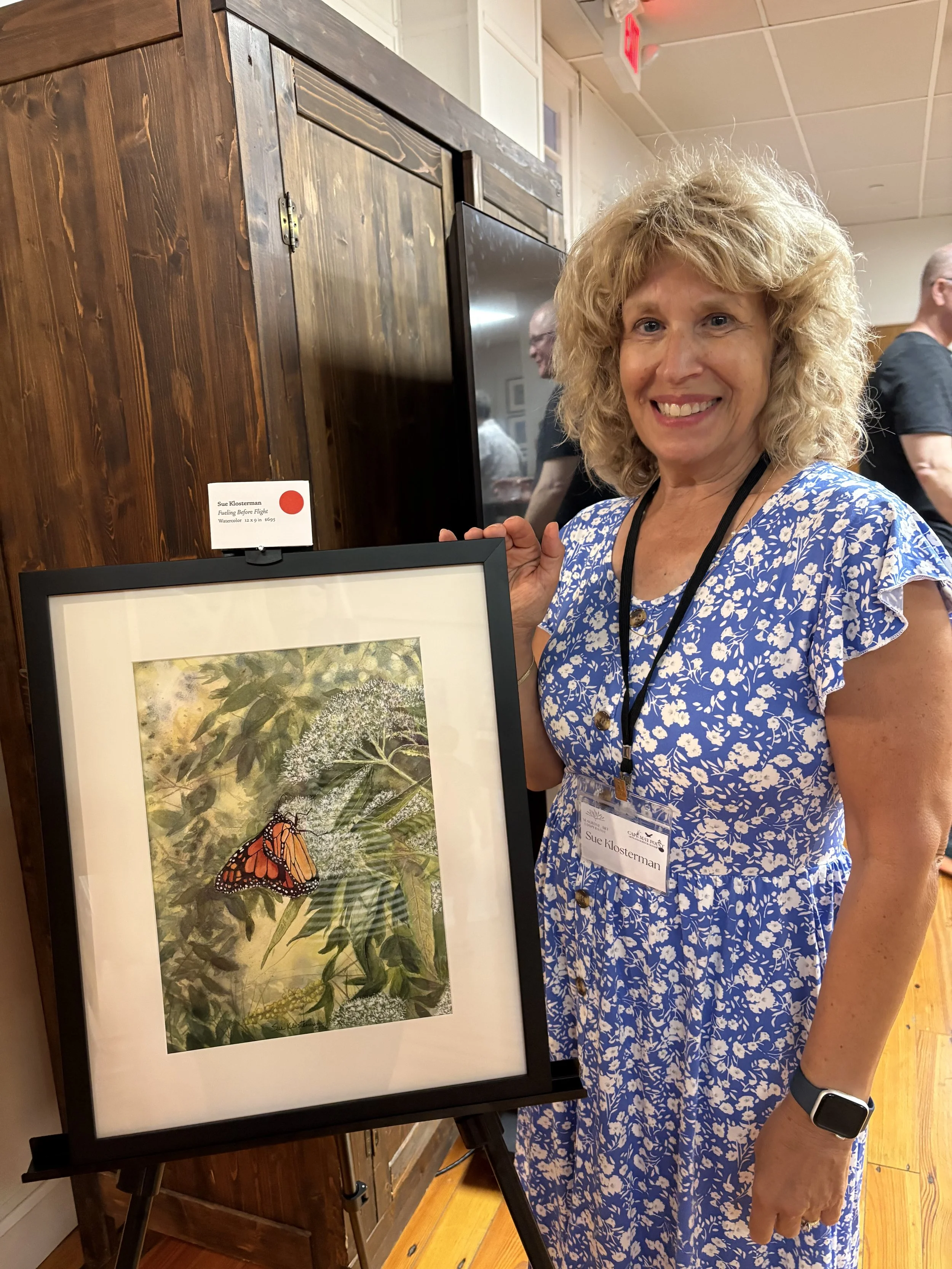 A woman with curly blonde hair and a blue floral dress is standing next to her artwork, which features a Monarch butterfly on a flower. She is smiling and holding the artwork stand, with a gallery setting in the background.