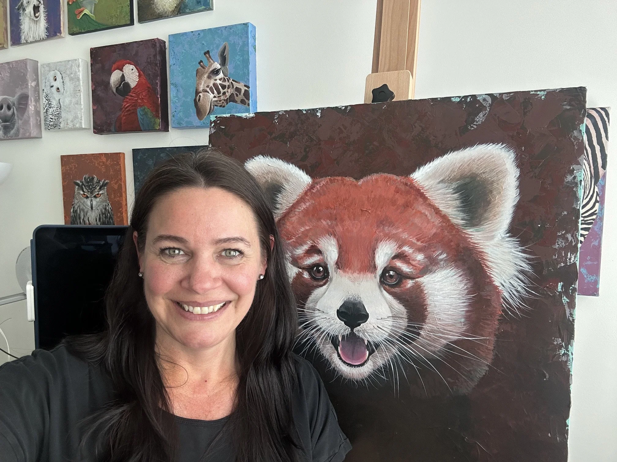 A woman with dark brown hair and light skin smiling while taking a selfie in front of colorful animal paintings including a large painting of a red panda and smaller paintings of a parrot, owl, and other animals on the wall behind her.