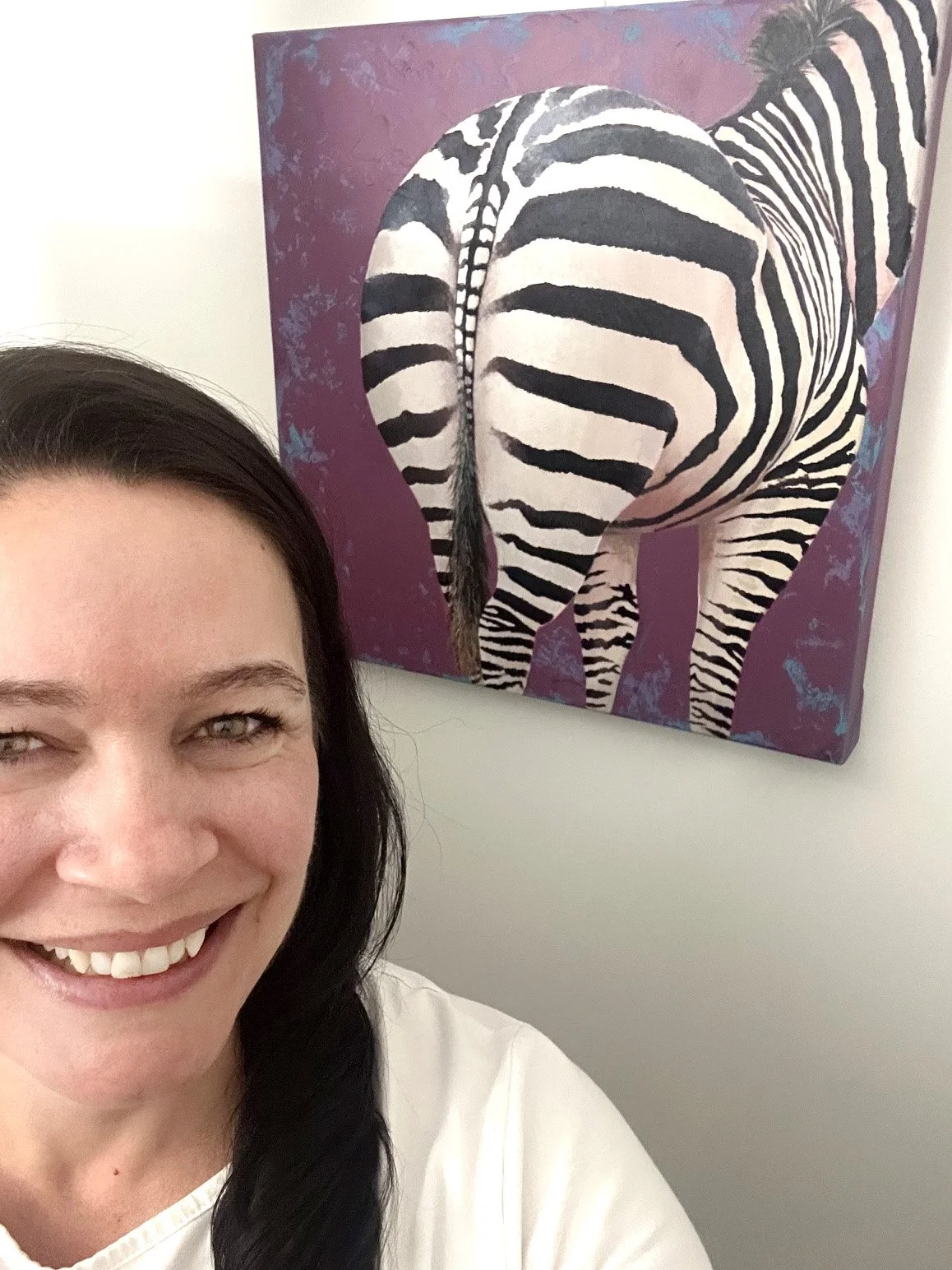 A woman with dark hair smiling in front of her unique painting of a zebra’s behind on a purple and blue background.