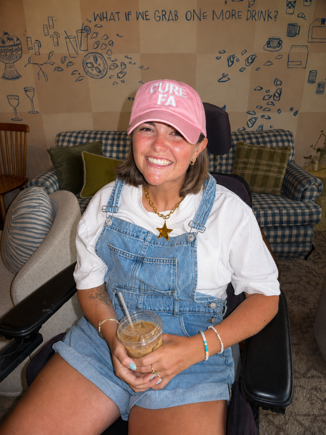 A young woman smiling and sitting in a wheelchair, wearing a pink baseball cap with 'CURE FA' embroidered on it, a white t-shirt under denim overalls, and holding iced coffee. The background has a wall with blue illustrations and text that reads, 'What if we grab one more drink?'