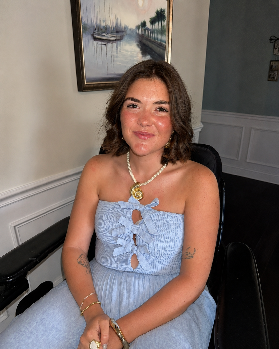 A young woman with shoulder-length brown hair, wearing a strapless light blue dress with bow details, seated in a black chair in a room with white wainscoting, a painting of boats and water on the wall, and decorative picture frames in the background.