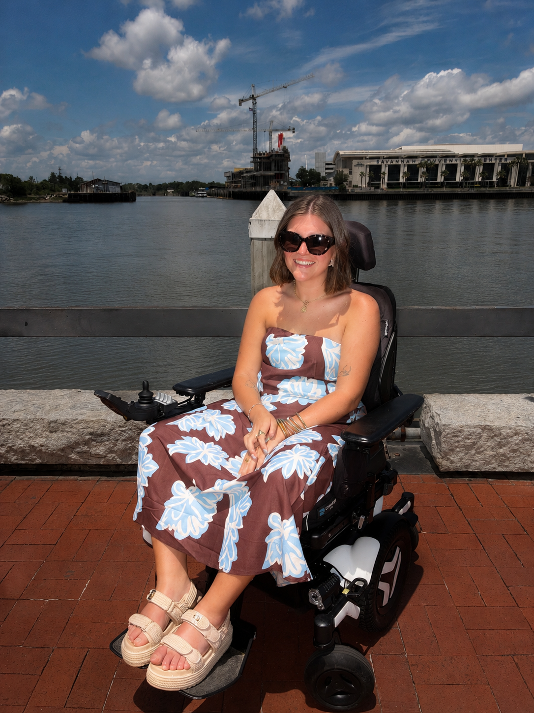 A young woman smiling and sitting in a motorized wheelchair on a brick walkway near a body of water, with construction cranes and buildings in the background under a partly cloudy sky.