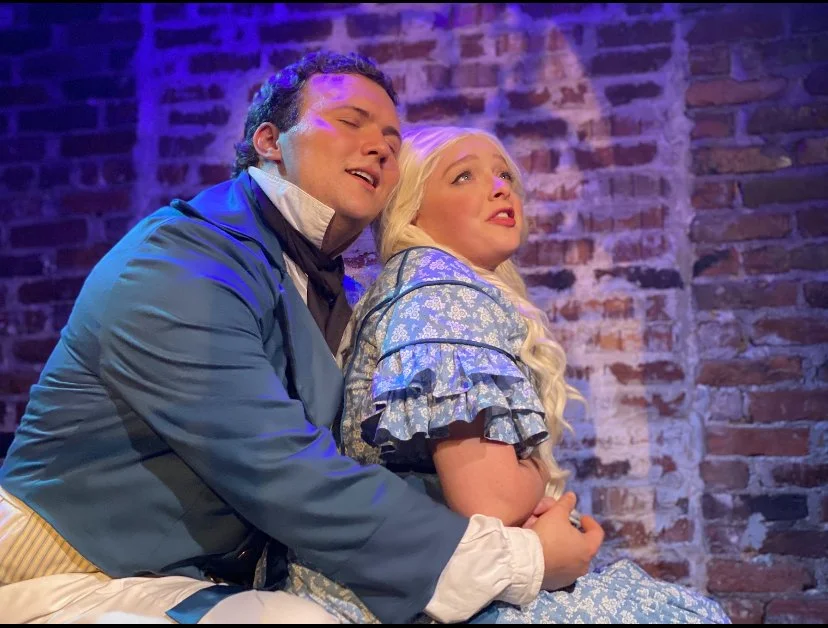 Two actors on stage, a man and a woman, performing in a theatrical play against an exposed brick wall background. The man, dressed in period costume with a blue coat, is sitting close to the woman and appears to be singing or speaking. The woman is w