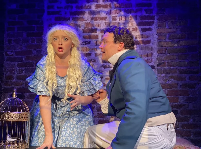 Two actors on a stage with a brick wall background, one woman with long blonde hair in a blue dress looking worried, and one man in blue and white costume, holding her hand.